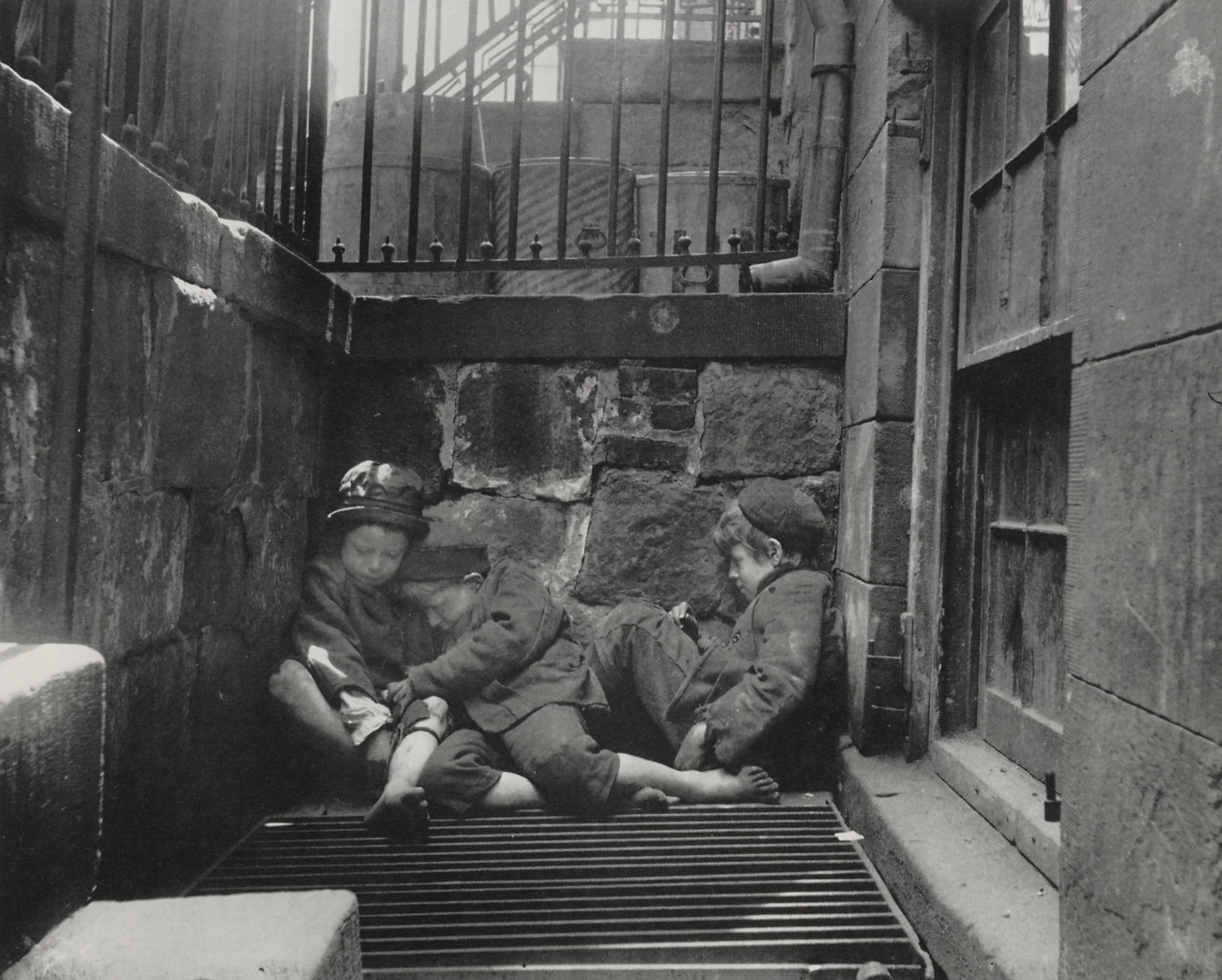 Three children in military-style clothing resting on a stone step in an alleyway, one leaning against a wall, two lying on their sides, surrounded by industrial and urban elements.