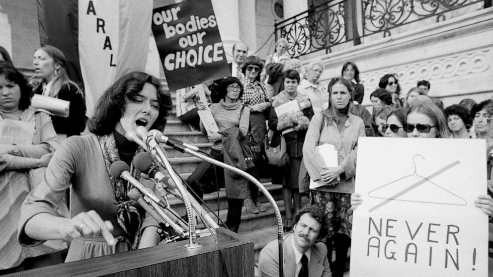 A woman speaking passionately at a microphone during a protest or rally, surrounded by a group of people holding signs that advocate for body autonomy and abortion rights, including a sign that says 'NEVER AGAIN!'