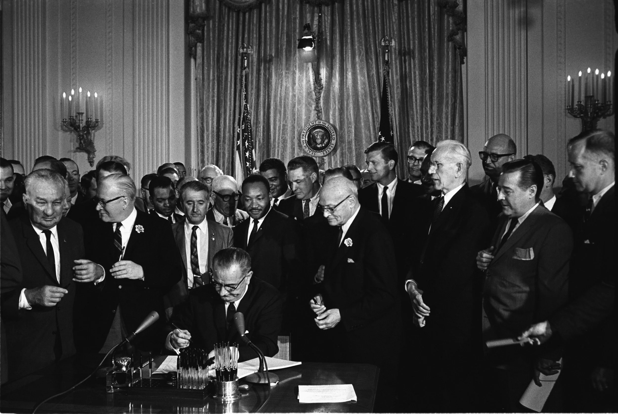 Black and white photo of a signing ceremony in a grand room with curtains, chandeliers, and the presidential seal. Several men in suits observe as one man signs a document on a table.