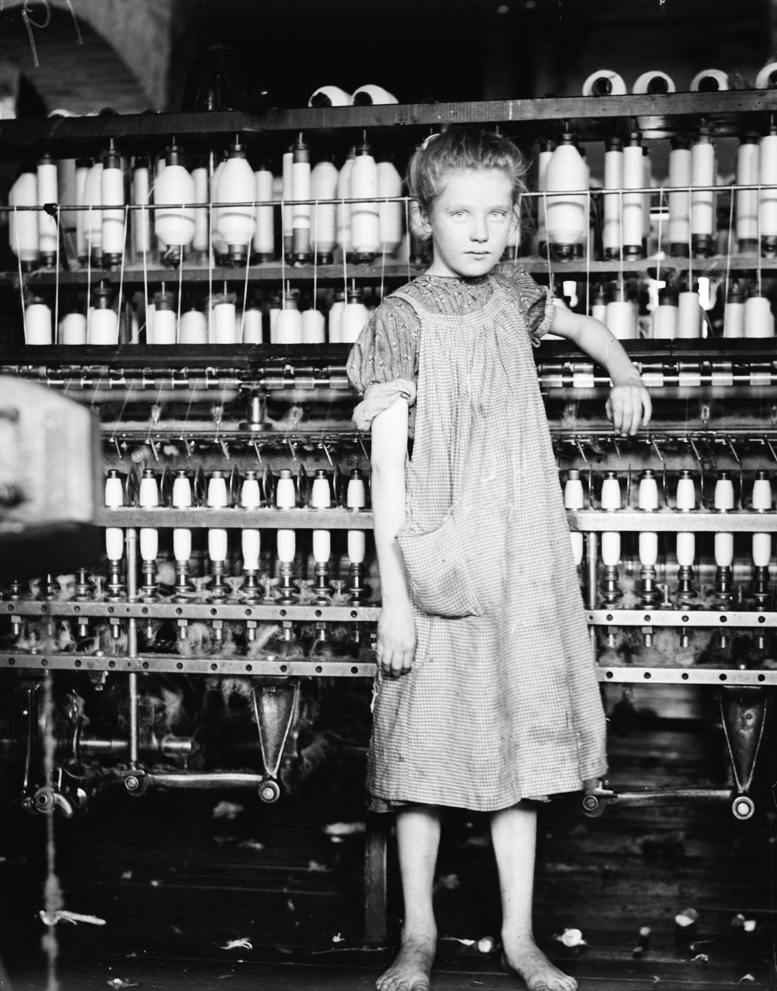 A young girl in a dress standing next to an industrial textile machine with spindles, likely in a textile mill.
