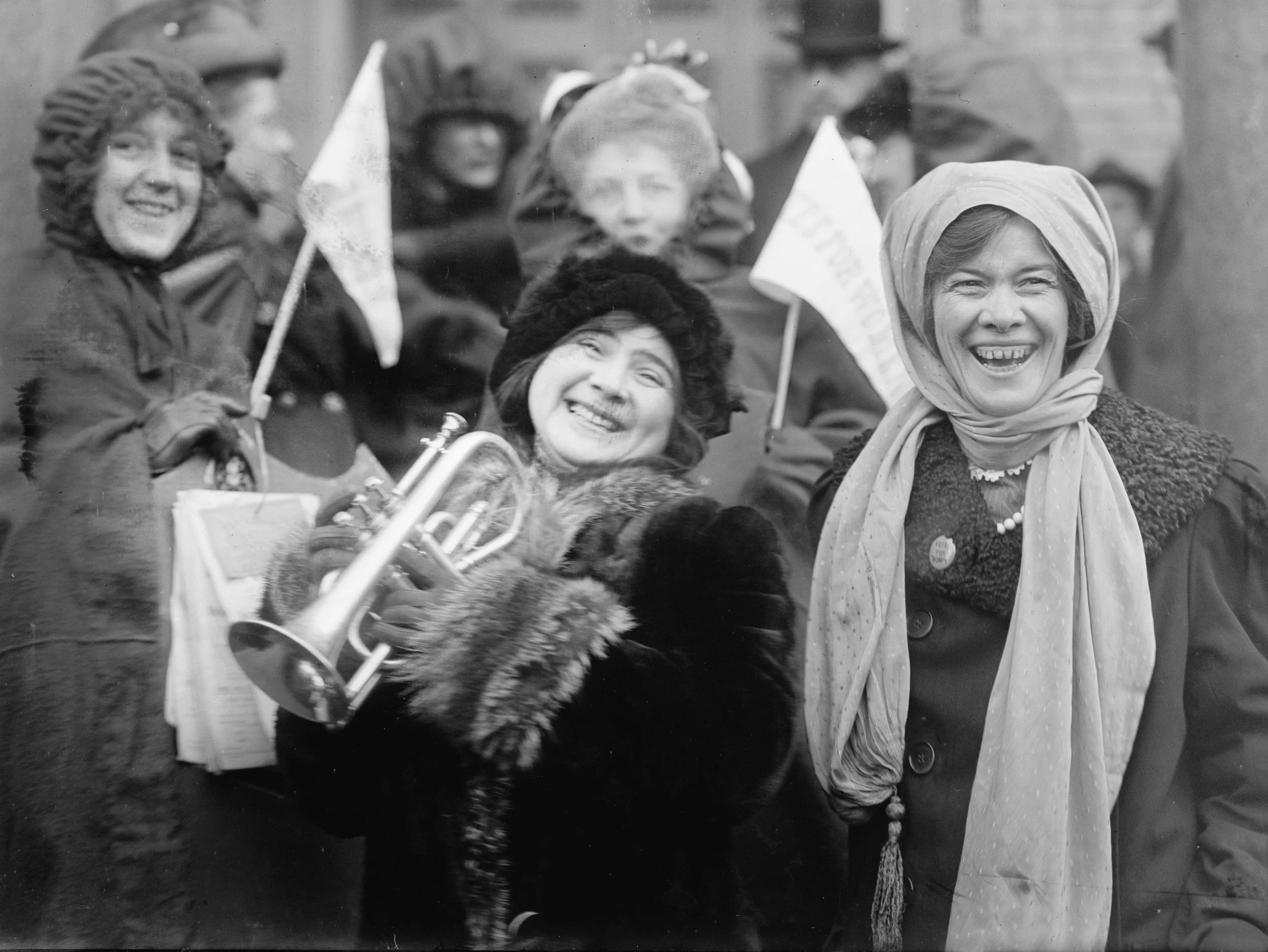 Women smiling, some holding small flags, in a gathering or parade, dressed warmly in coats and scarves, black and white photo.
