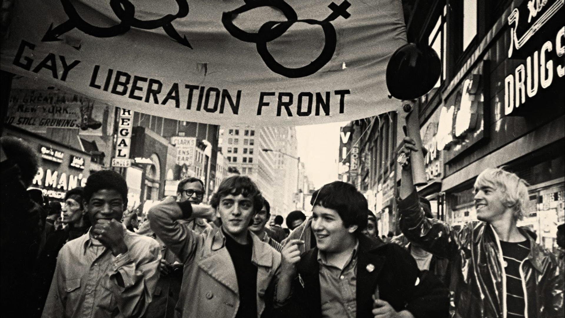 A black and white photo of a group of people marching during a Pride parade in New York City, holding a sign that reads 'Gay Liberation Front,' with buildings and signs visible in the background.