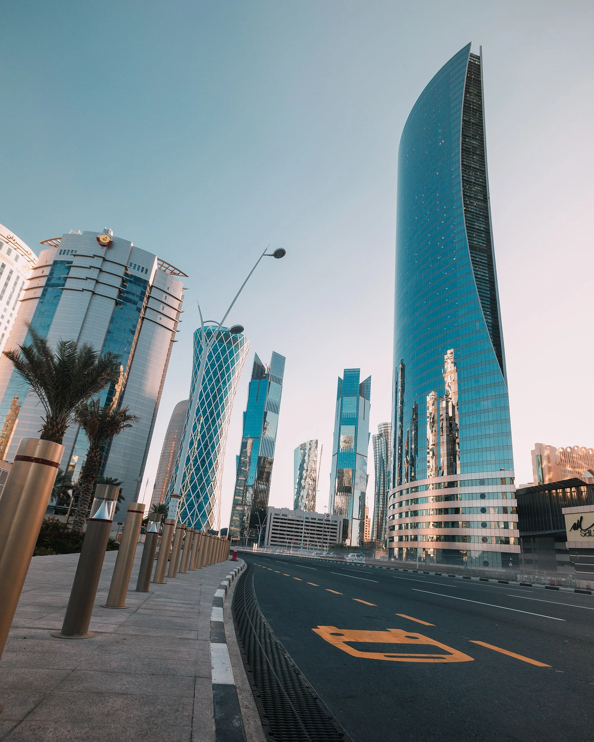 Cityscape of modern skyscrapers with glass facades, a curved approaching road marked with a yellow symbol, and palm trees along the sidewalk.