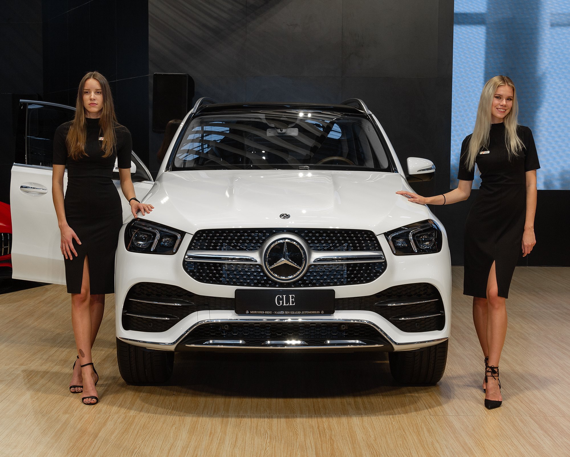 Two women in black dresses standing next to a white Mercedes-Benz GLE SUV at an auto show.