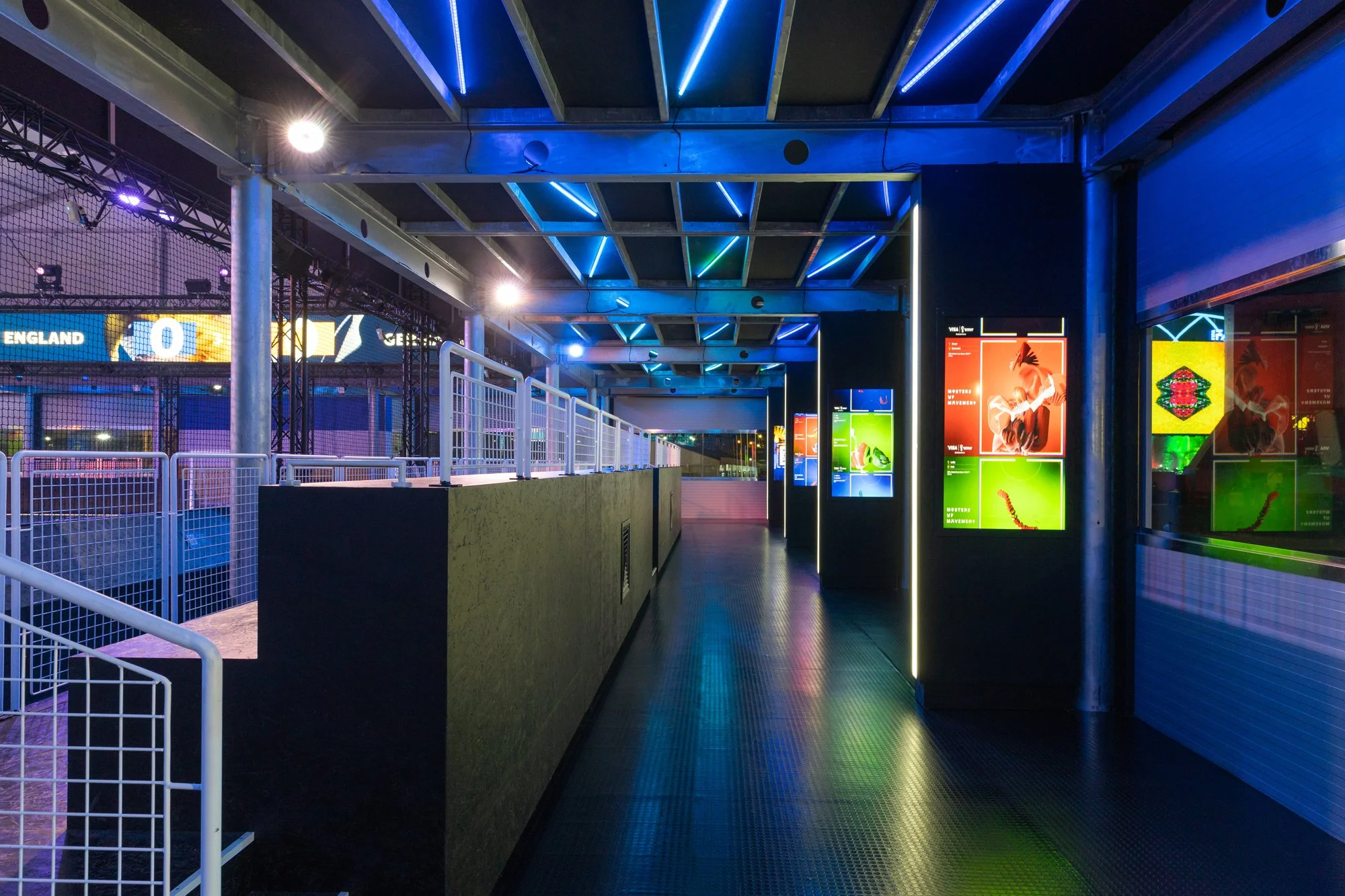 Empty modern sports stadium corridor with colorful illuminated advertisements and a railing on the left, illuminated with blue and green LED strip lights on the ceiling.