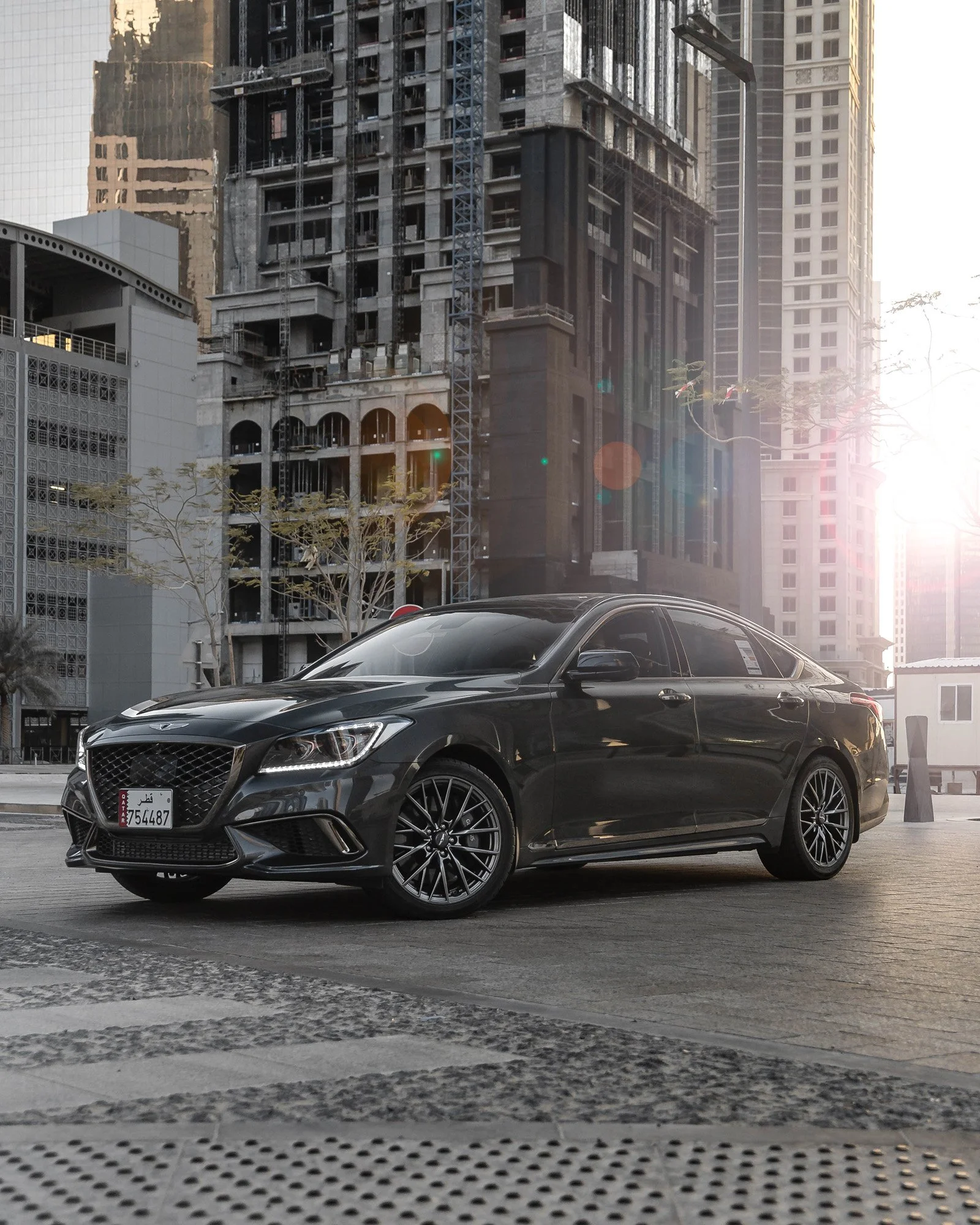 Genesis G70 parked at West Bay Dafna with tall buildings and a construction site in the background, sunlight shining from the right in Doha, Qatar.