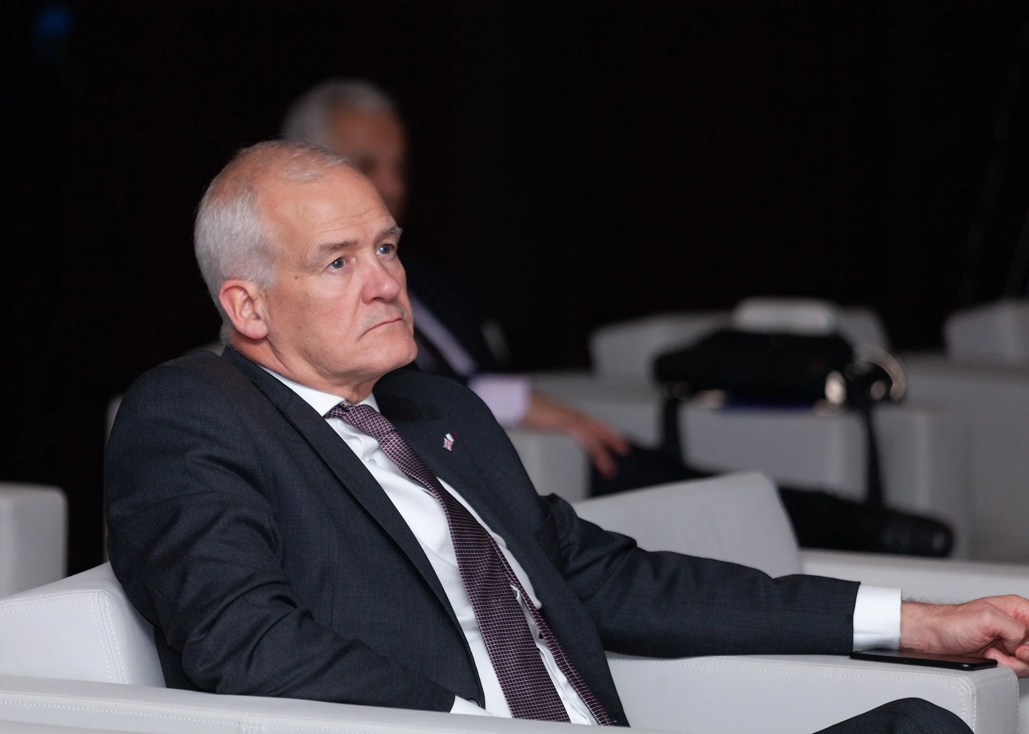 An elderly man with gray hair, dressed in a dark suit with a white shirt and patterned tie, sitting on a white couch in a dark room, attentively listening or watching.