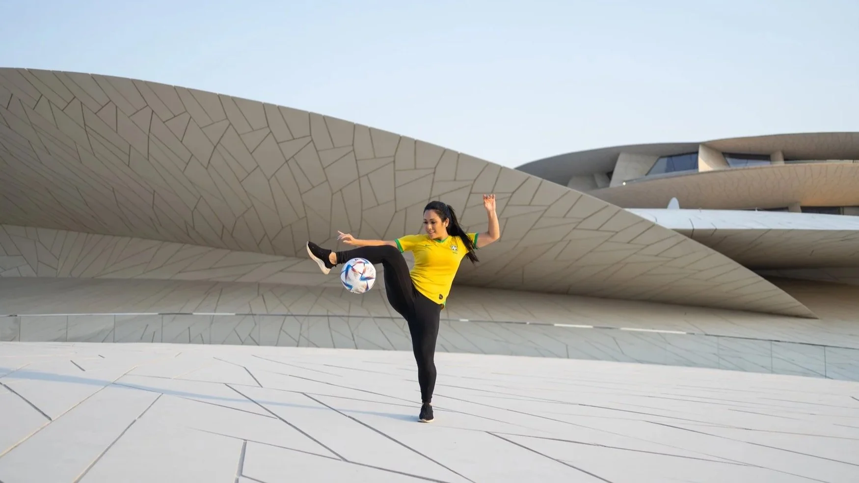 Brazilian freestyler Raquel Benetti with Qatar Creates a yellow sports jersey and black pants is performing a soccer trick with a ball in front of National Museum of Qatar in Doha, Qatar.