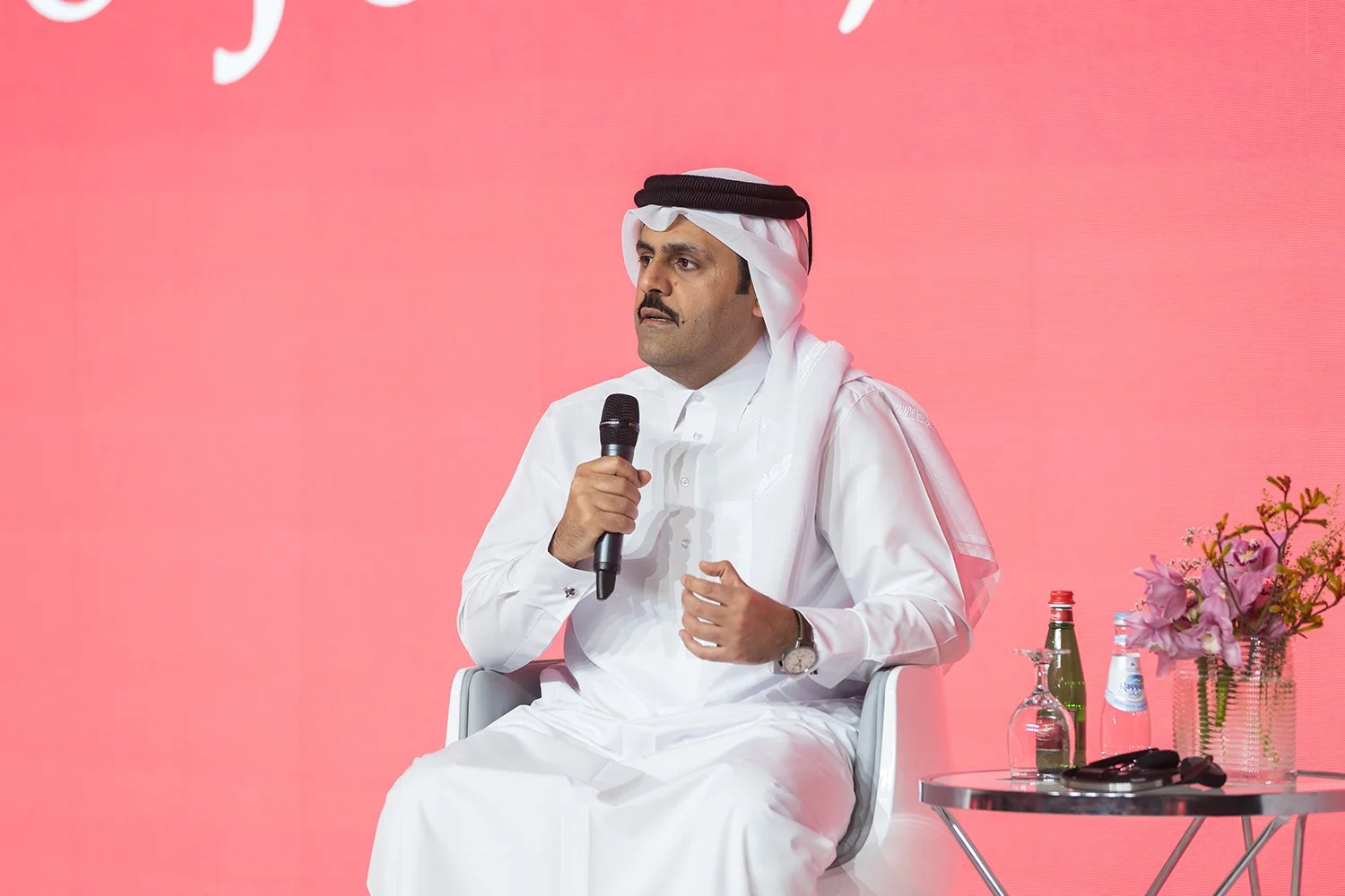 A man in traditional Middle Eastern attire, including a white thobe and a head covering, sitting on a white chair holding a microphone. There is a small table with water bottles, a glass, and a bouquet of pink flowers beside him, set against a pink b