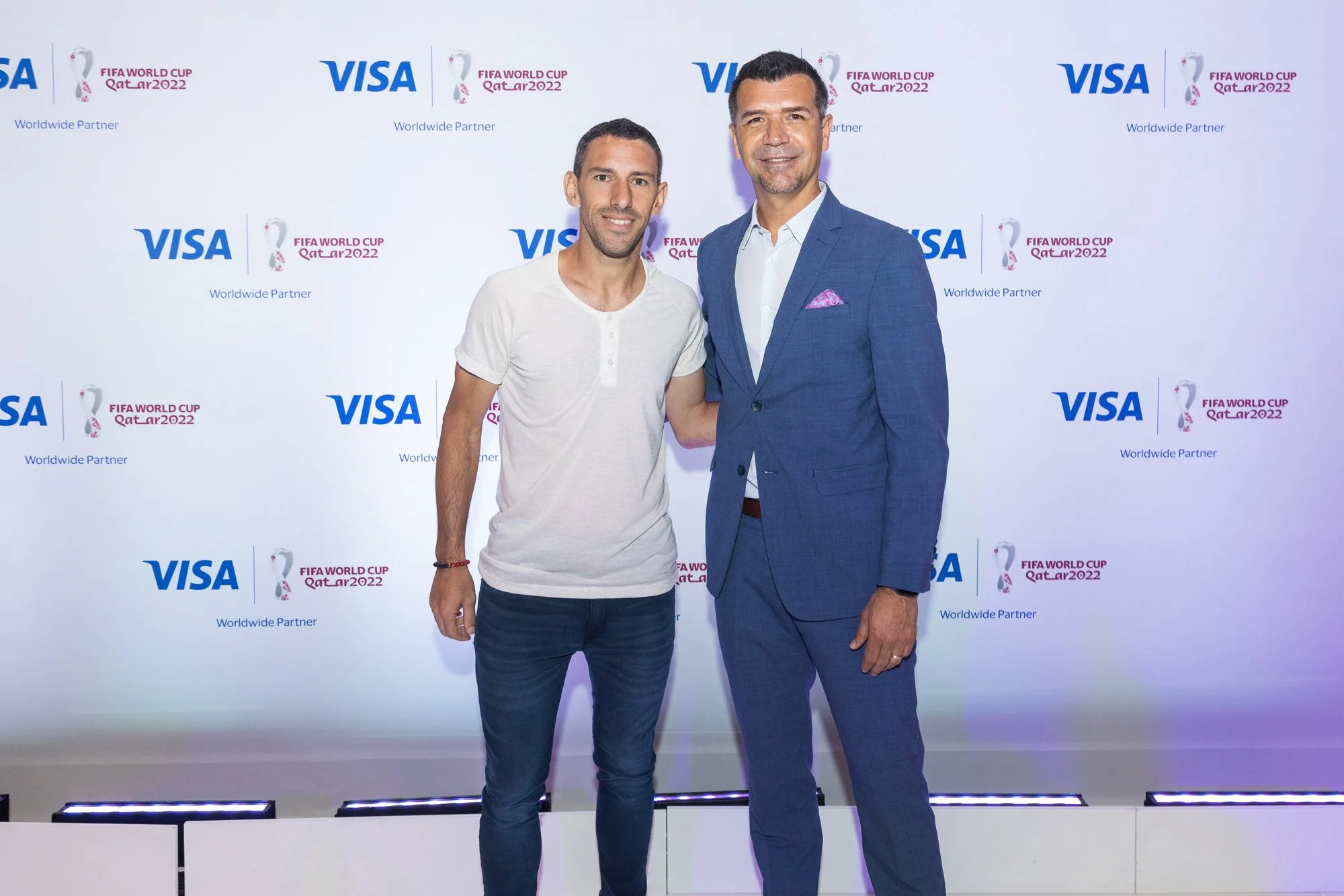 Two men standing in front of a step-and-repeat banner with logos of FIFA World Cup Qatar 2022, VISA, and a FIFA World Cup icon. One man is wearing a white t-shirt and jeans, and the other is dressed in a blue suit with a white shirt and pink pocket s