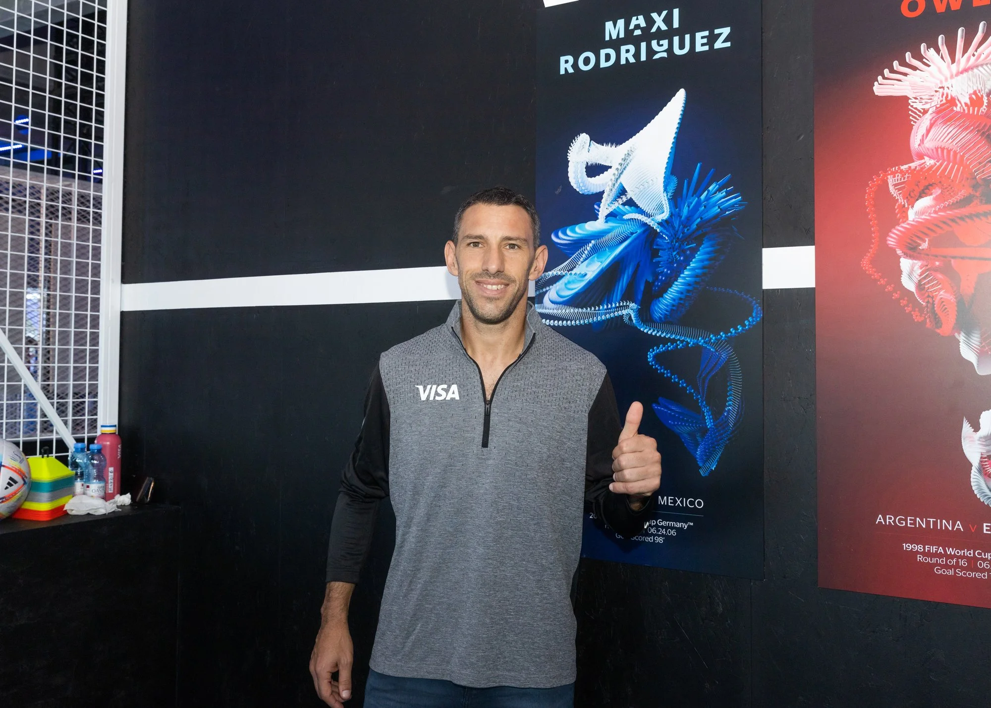 A man giving a thumbs-up gesture at a sports event, standing in front of a large poster featuring a blue abstract digital illustration of a musical instrument and the text 'Maxi Rodriguez' and 'Mexico'.