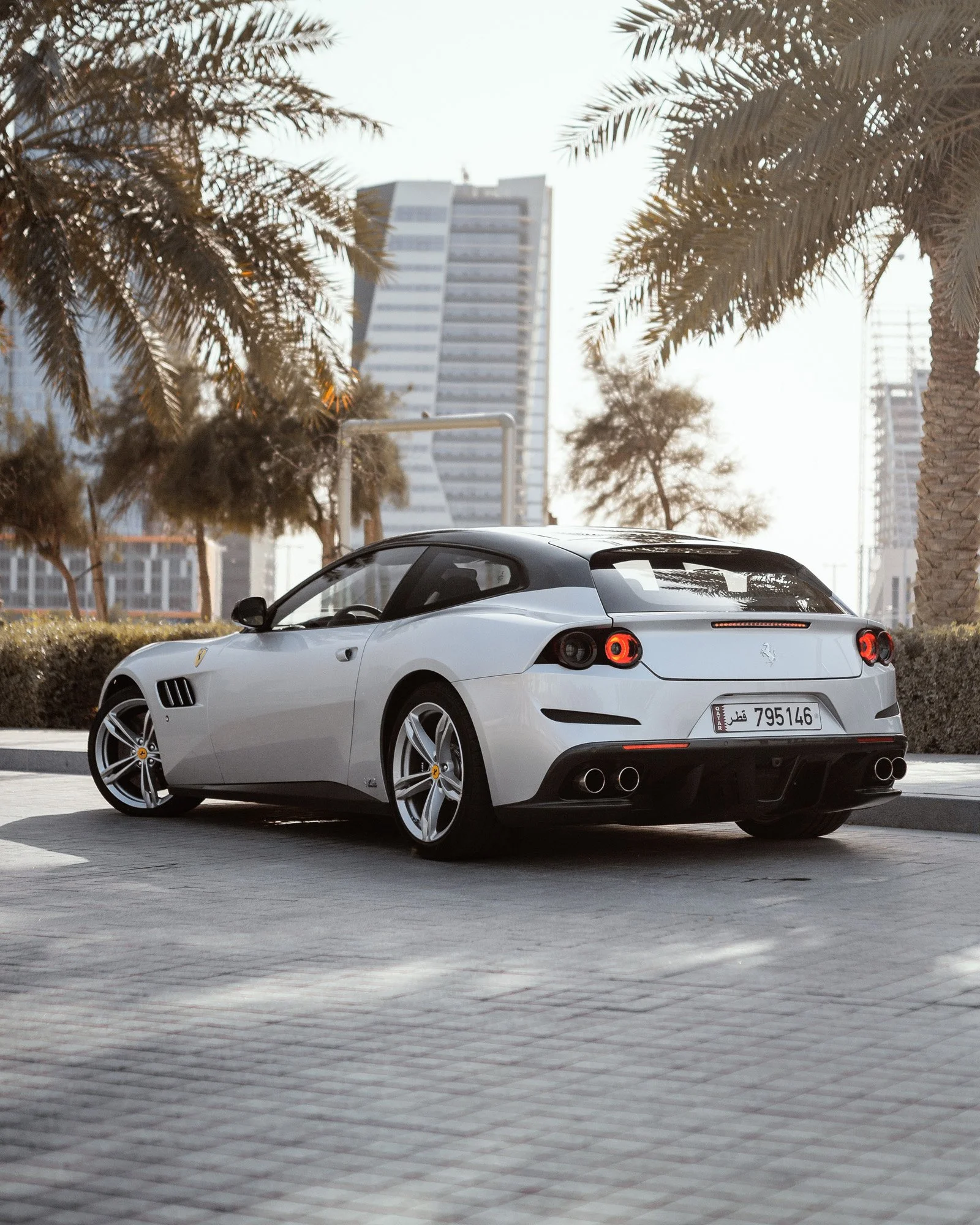 A white Ferrari sports car parked on a city street with palm trees and tall buildings in the background during daylight.