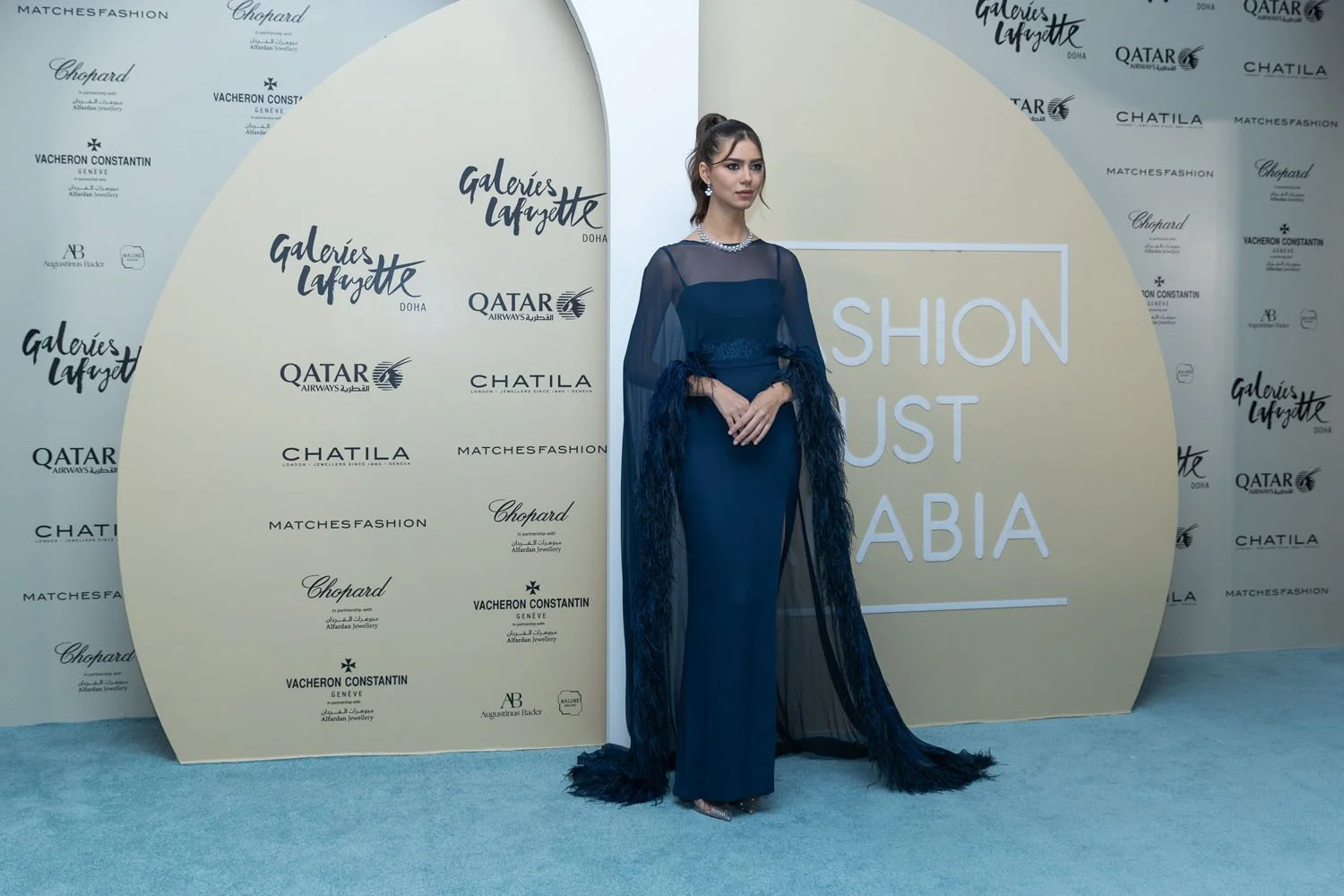 A woman in an elegant blue gown with sheer overlays and feathered sleeves stands on a blue carpet at a fashion event. Behind her is a backdrop with logos, including "Galeries Lafayette Doha," "Qatar Airways," "Chopard," "Chatila," and "MatchesFashion
