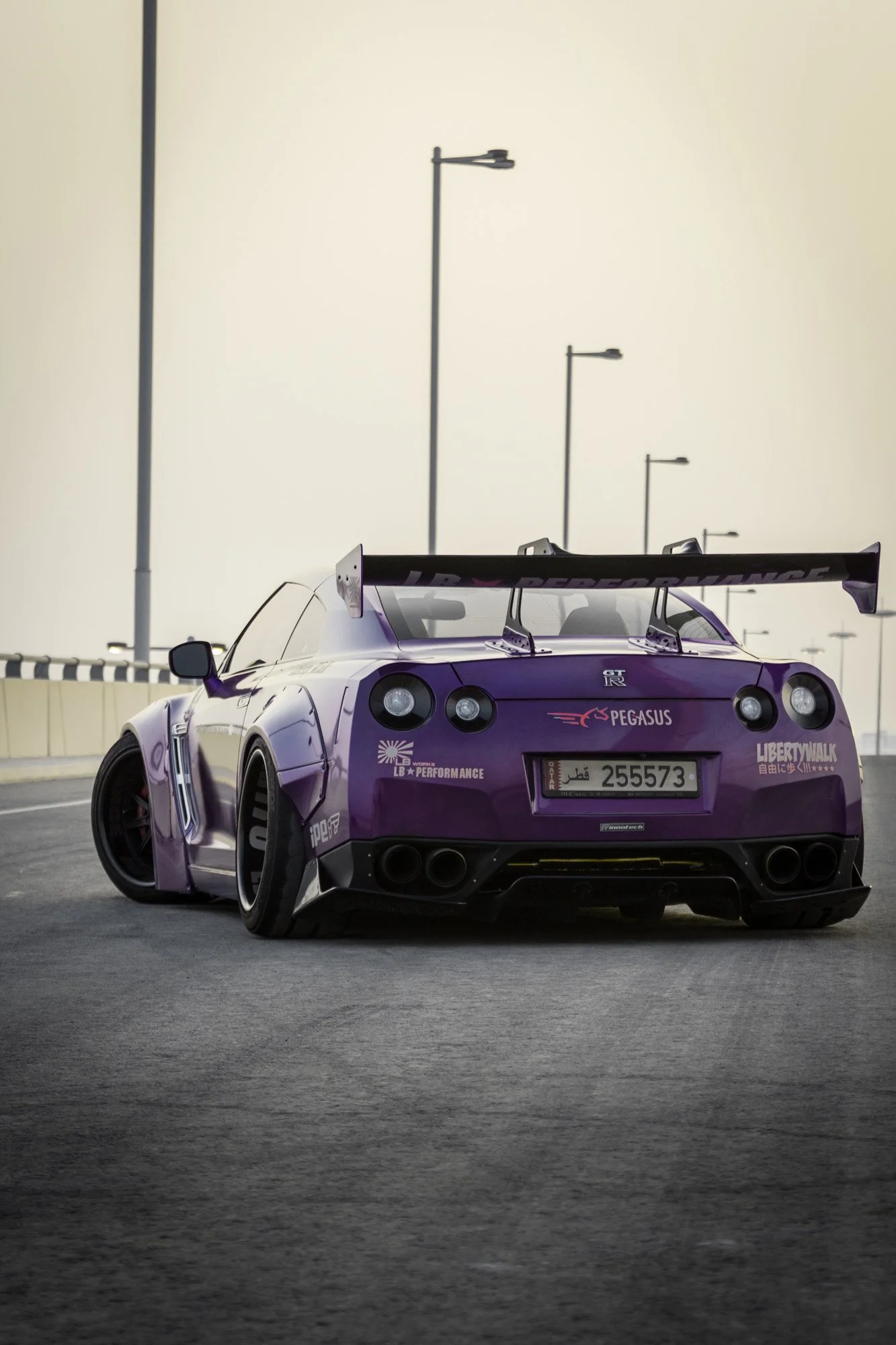 A purple race car with a large rear wing on an empty road during daytime, some streetlights in the background.