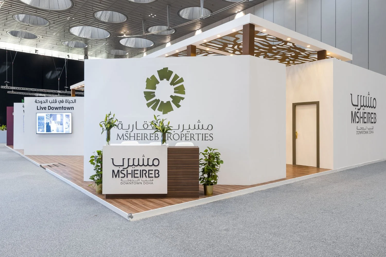 Trade show booth for MSHEIREB Properties in Doha, featuring a white and brown color scheme, potted plants, and signage in Arabic and English, with the MSHEIREB logo on the wall.