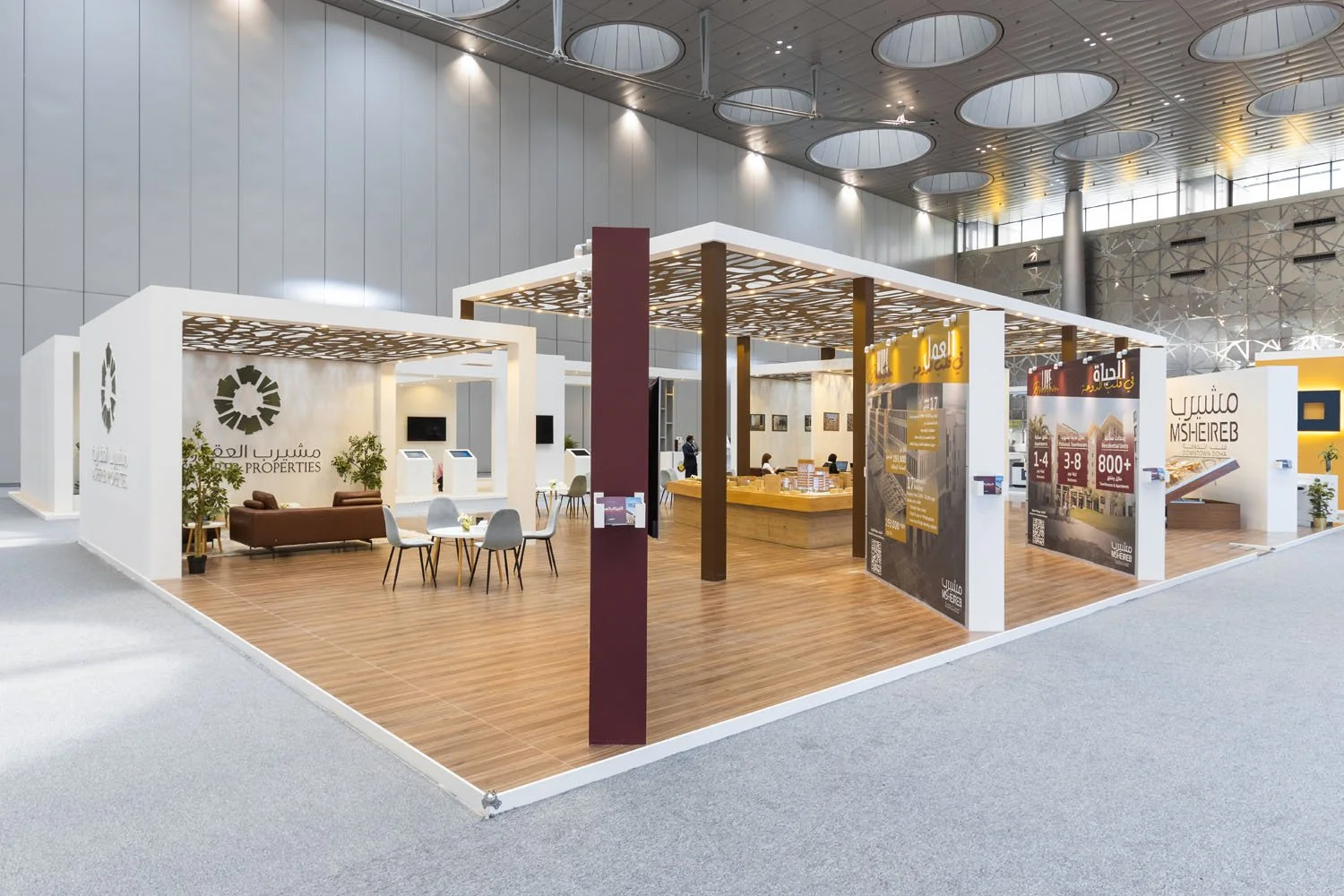 Exhibition booth with a modern design, featuring sections labeled 'Properties' and 'Ms. Eireb,' with seating areas, informational displays, and a reception counter inside an open gallery space.