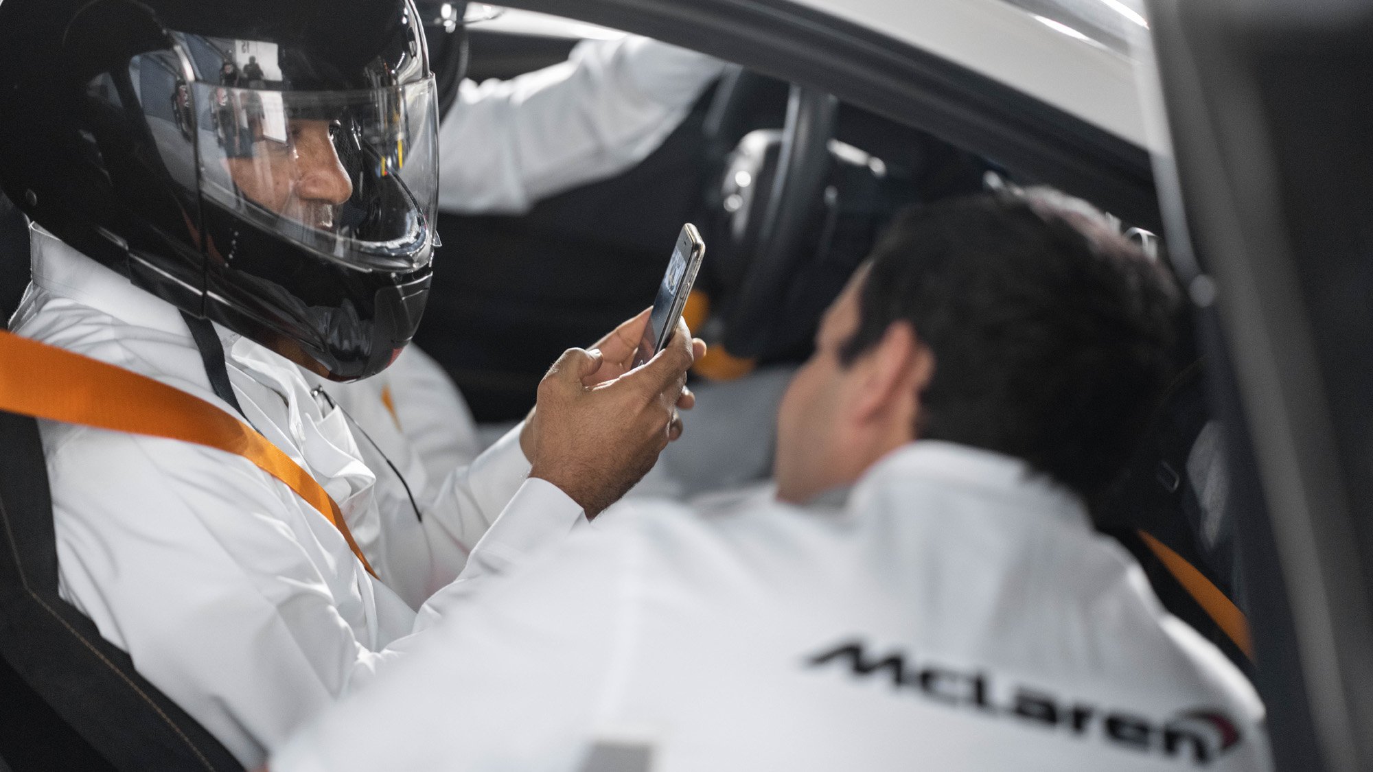 Two race car drivers in white racing suits with McLaren logos sitting inside a race car; one is wearing a black helmet and looking at his phone, while the other is resting with their head back.
