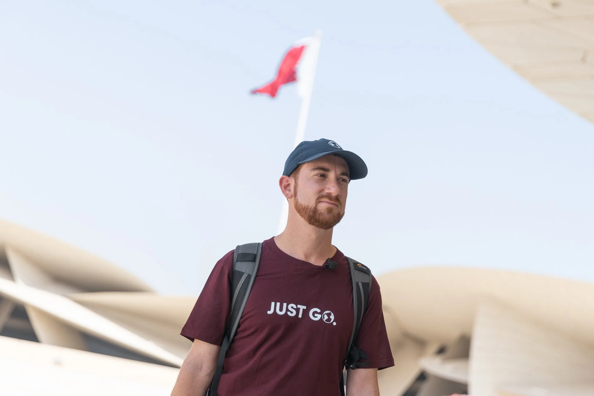 Drew Binsky wearing black cap, a maroon T-shirt with 'JUST GO.' printed on it, and a backpack, standing outdoors during daytime with National Museum of Qatar and Qatari flag in the background.