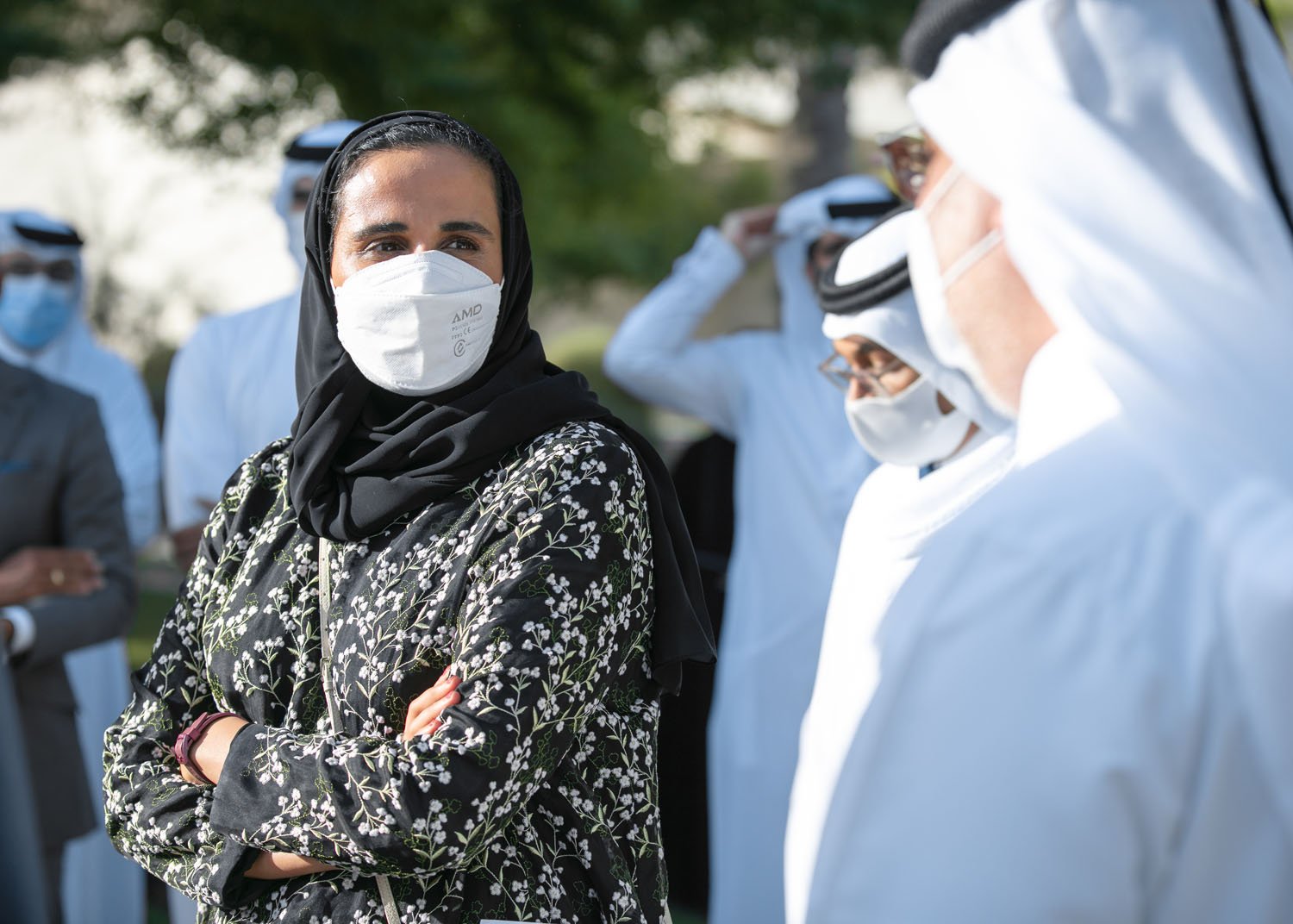 A woman wearing a black hijab and face mask standing outdoors with her arms crossed, surrounded by men in white traditional Arab attire and face masks, in a green outdoor setting.