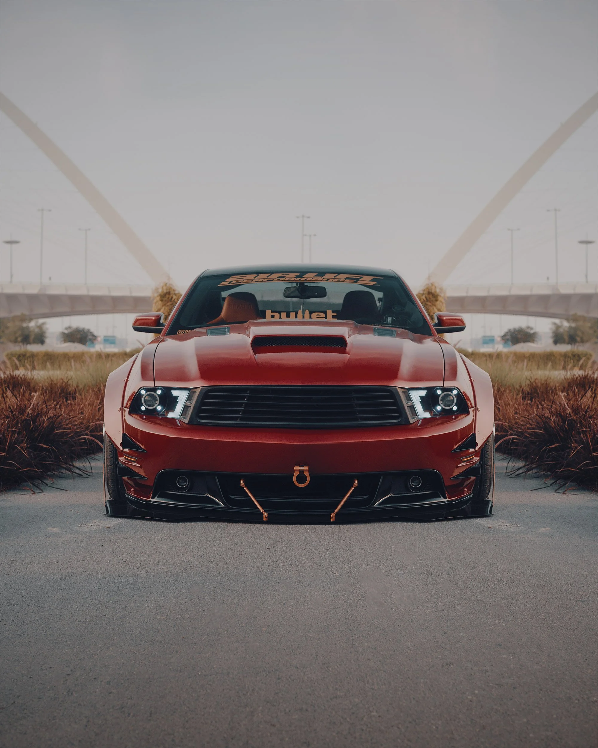 Red Ford Mustang GT with black accents parked on a paved road, low stance, under a bridge, with a clear sky at Lusail in Doha, Qatar.