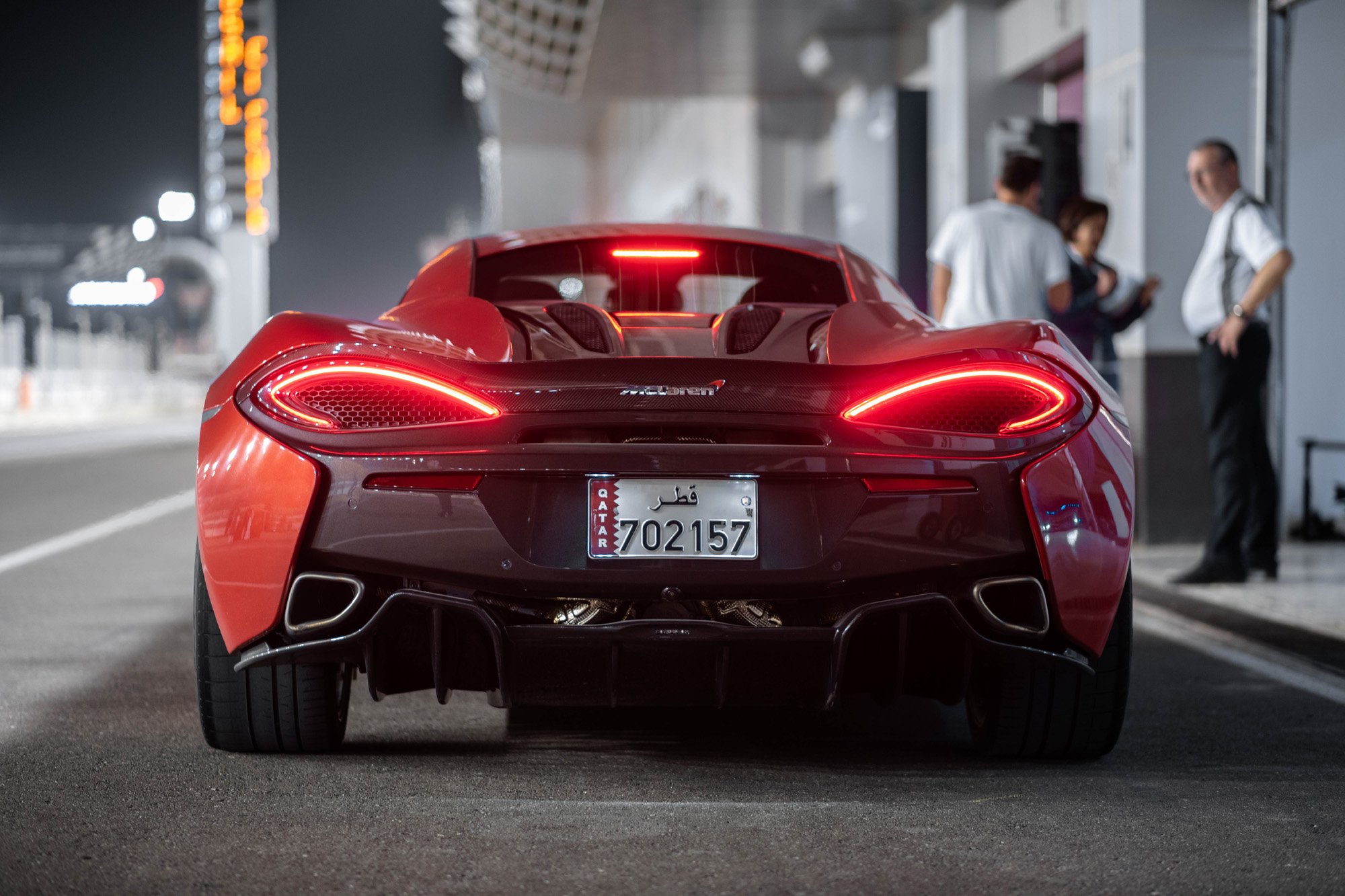 Red McLaren supercar at a Lusail International Circuit in Doha, Qatar, viewed from the rear, with a Qatar license plate, and people in the background.