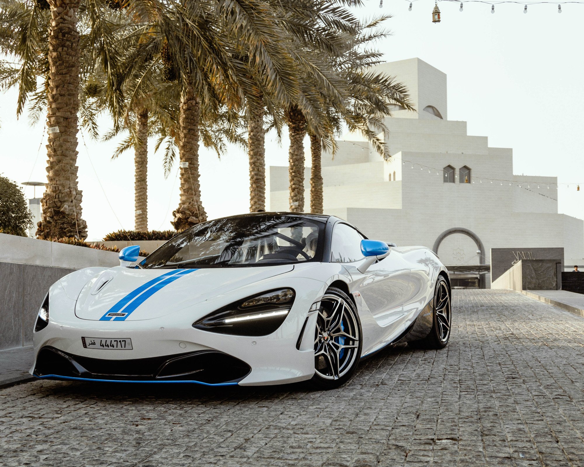 A Mclaren with blue accents parked on a cobblestone street, with palm trees and a modern white building in the background at Museum of Islamic Art in Doha, Qatar.