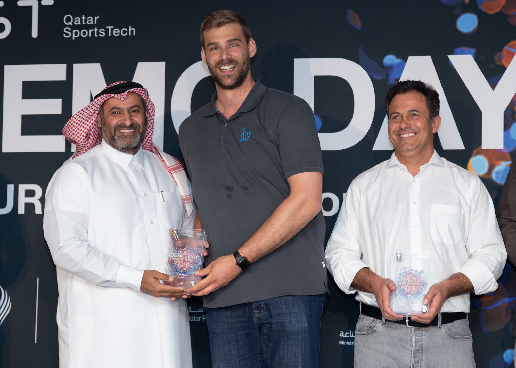 Three men pose for a photo at a Qatar SportsTech event, with two of them holding glass awards. The background features the text 'DEMO DAY'.