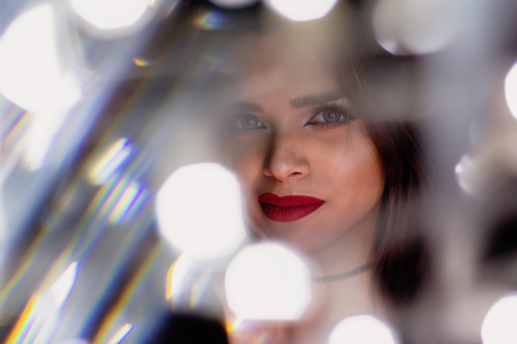 Model Marie D'Souza with dark hair and red lipstick looking through a reflective surface or mirror with bright out-of-focus lights creating a bokeh effect at Doha, Qatar.
