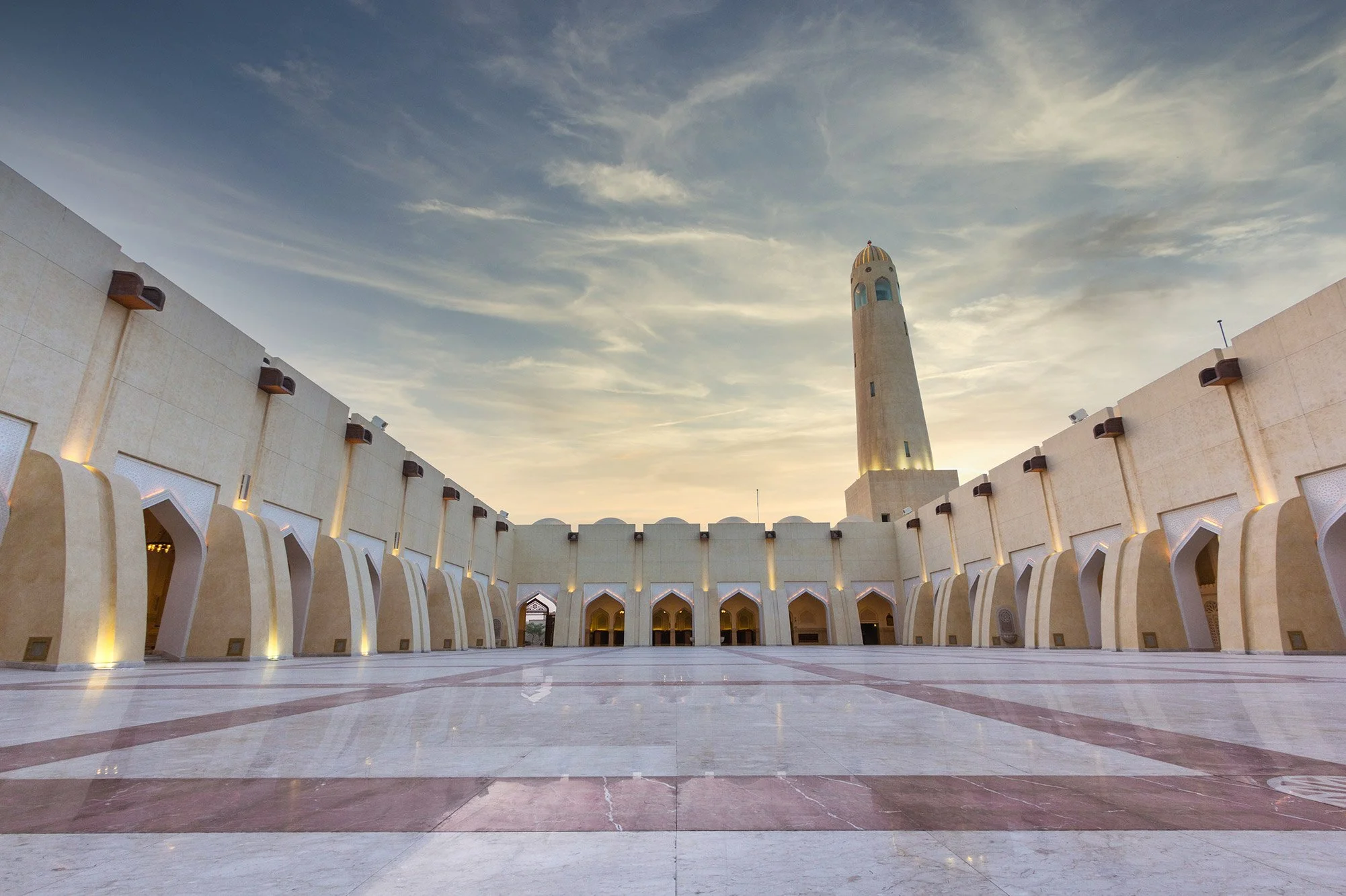 Empty courtyard with white marble flooring and beige walls, featuring a tall minaret in the background under a partly cloudy sky at sunset.