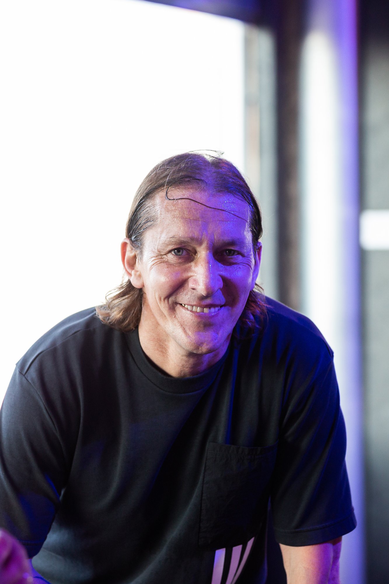 A man with medium-length brown hair, smiling, wearing a black t-shirt, in a brightly lit indoor setting with white and purple lighting.