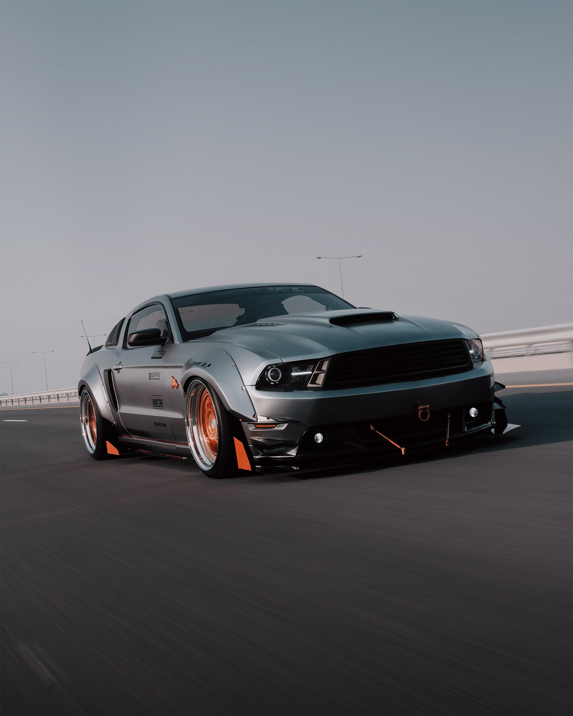 A sleek, Ford Mustang GT car with custom wide wheels, racing decals, and aerodynamic modifications moves on an empty Orbital highway at sunset in Doha, Qatar.