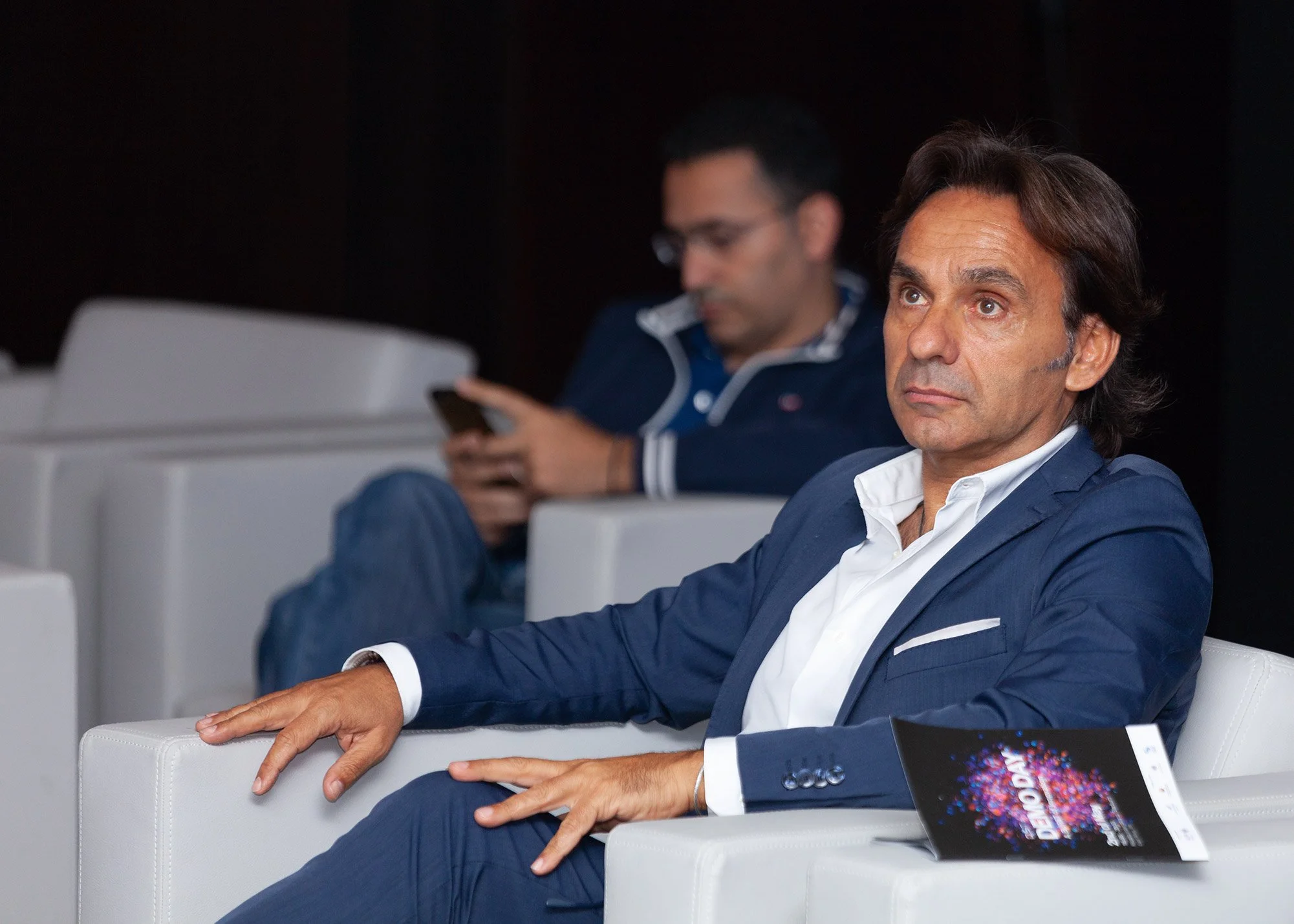 Two men in business suits sitting in white chairs, one in the foreground looking attentive, the other in the background using a phone, with a black background.