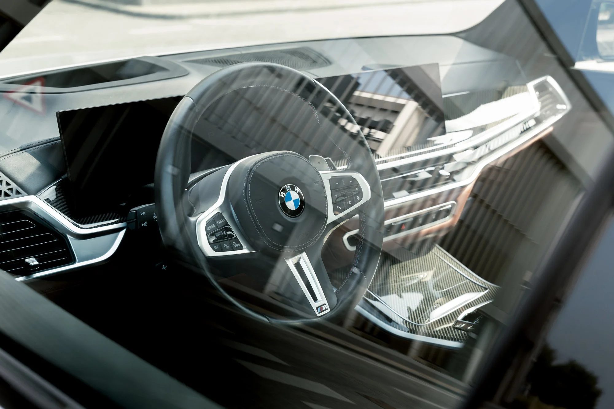 Interior view of a BMW X5 M60i vehicle, showing a steering wheel with BMW logo, digital instrument cluster, and modern dashboard with carbon fiber accents, seen through the windshield. Photography by Talha Belal
