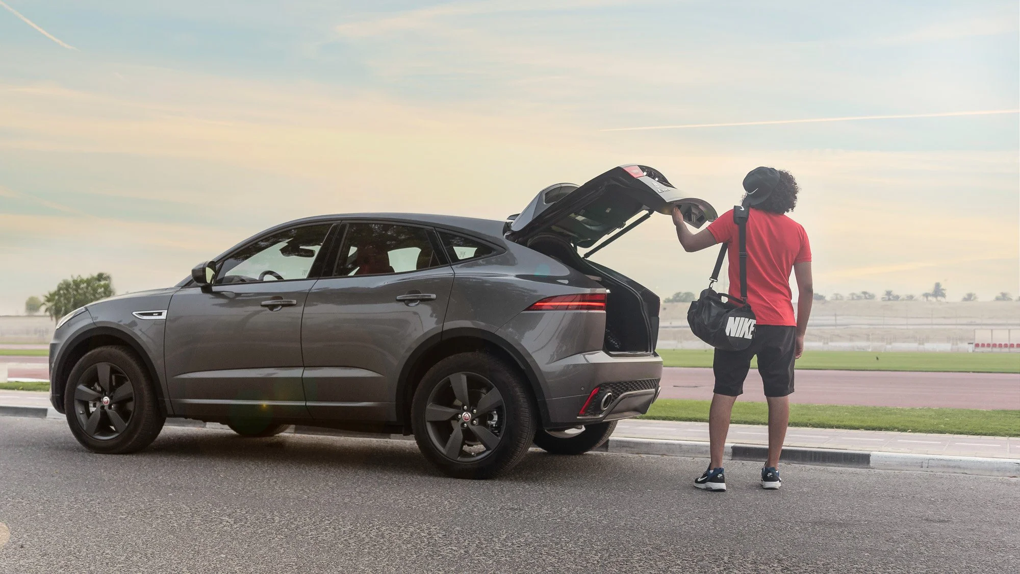 A young man in a red t-shirt and black shorts, wearing a black Nike bag and headphones, stands next to a grey SUV on a road with an open trunk during sunset or sunrise.