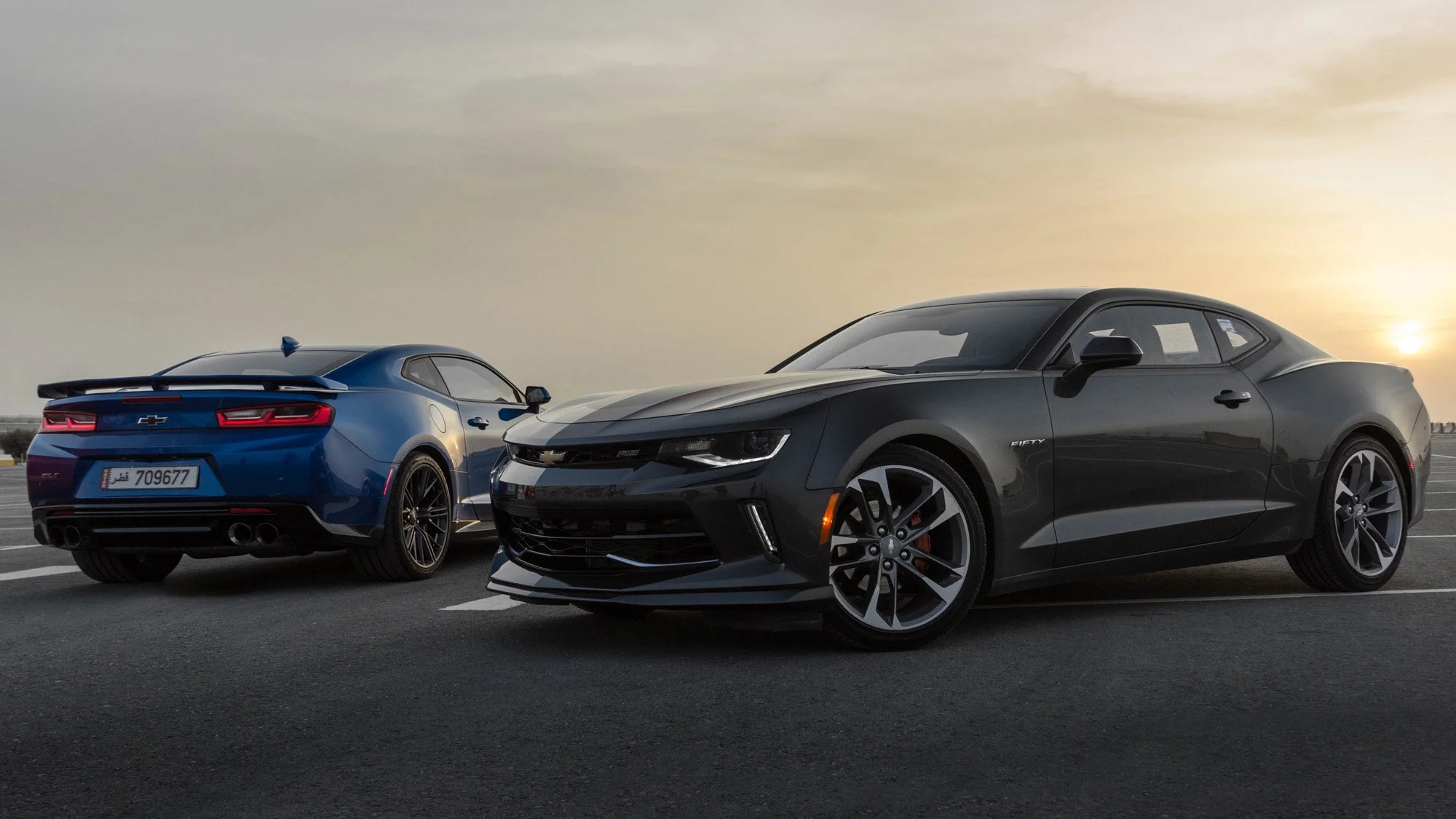 Two Chevrolet Camaro from Al Jaidah Motors parked on an empty asphalt surface at sunset, with a yellowish sky in the background at Doha, Qatar.