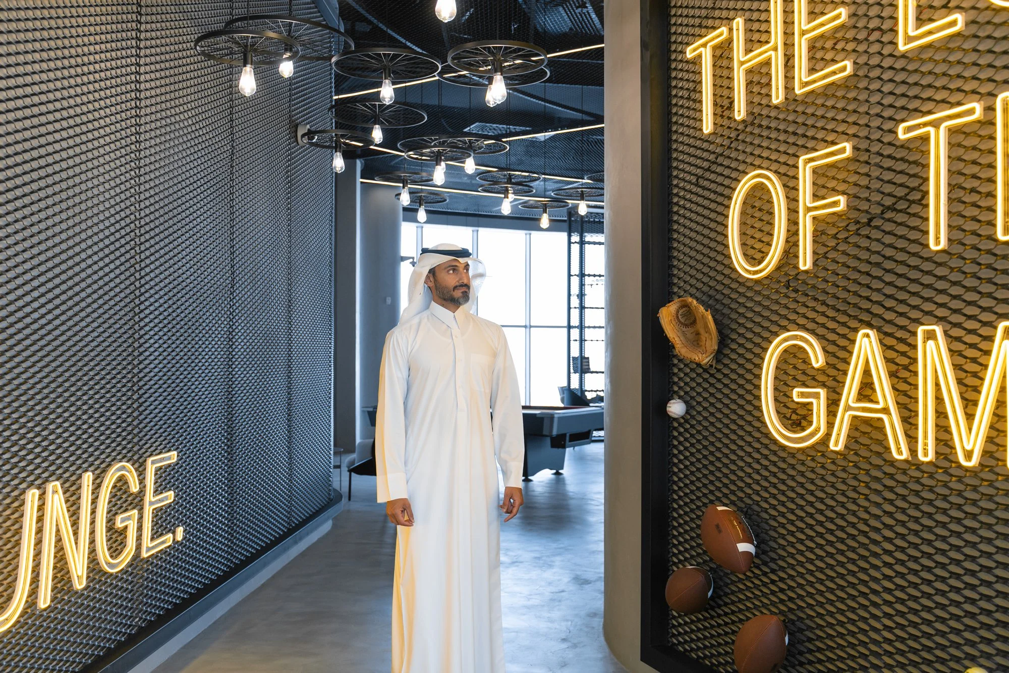 A man in traditional Middle Eastern attire stands inside a modern building with geometric light fixtures and neon signs reading 'The of to GAV' on a textured wall.