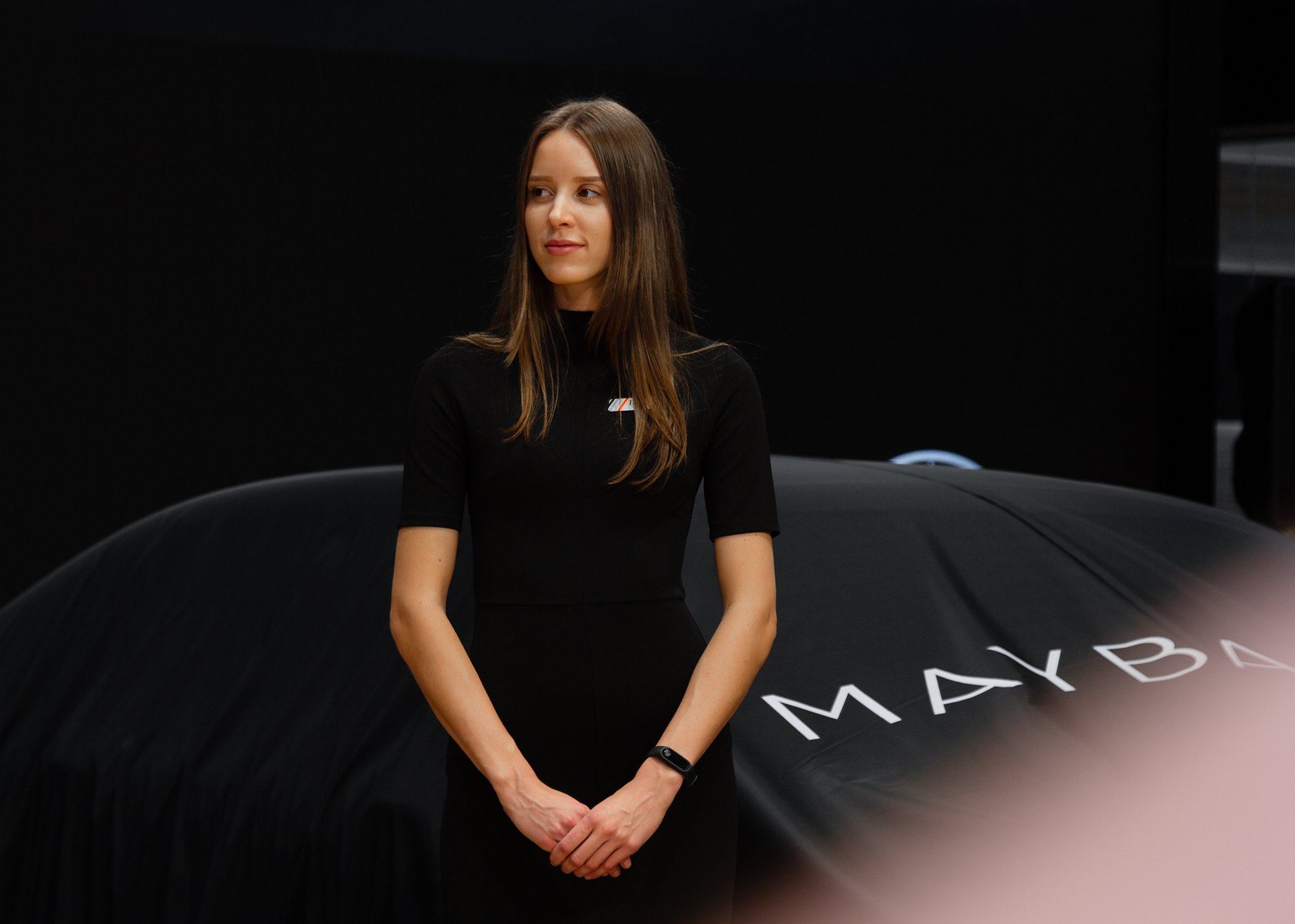 A woman with long brown hair in a black dress stands next to a covered car with the word "MAYBA" visible on the cover.