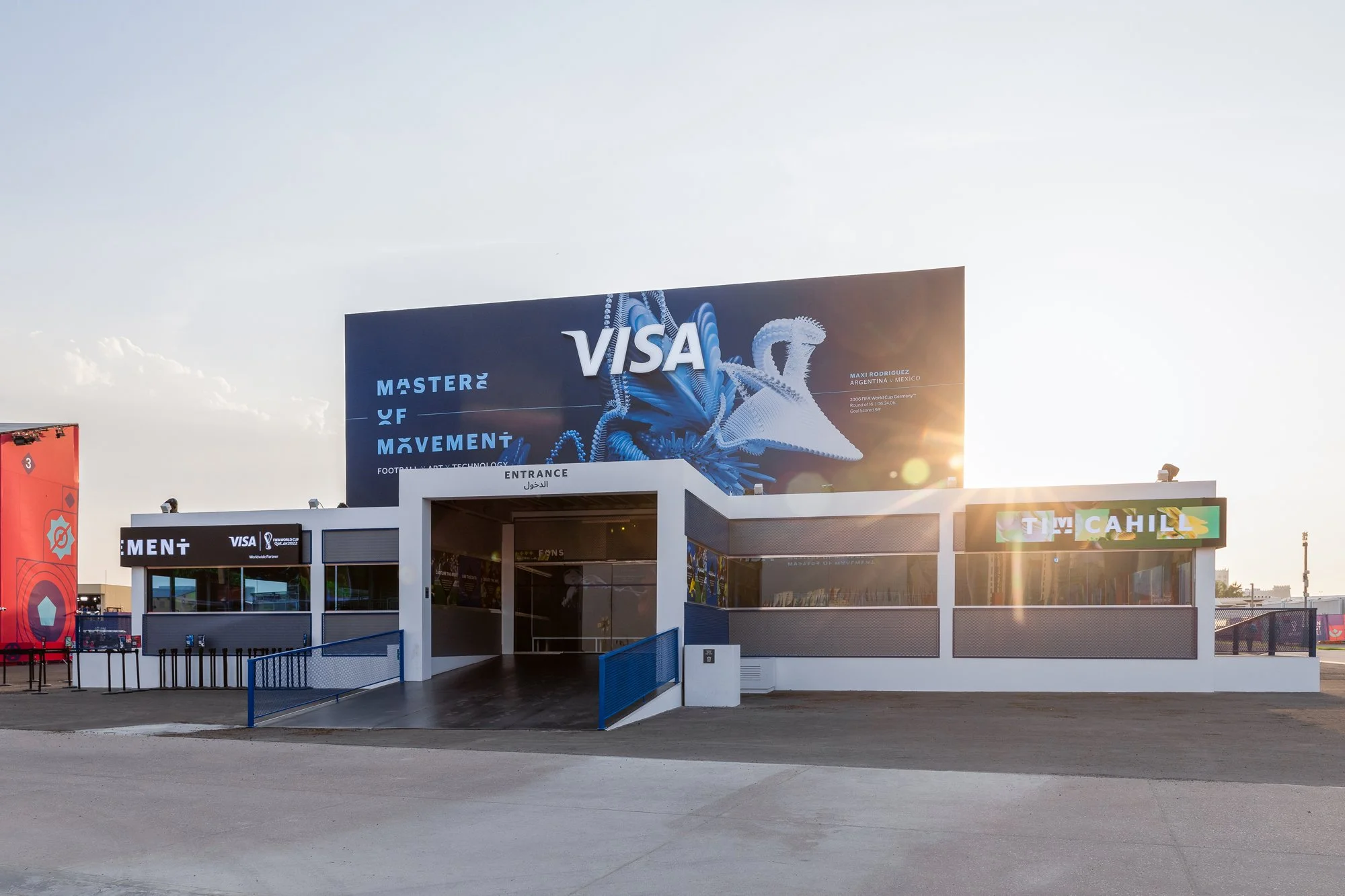 Empty indoor event venue with digital screens and a large billboard displaying the Visa logo and promotional images, with a clear sky and sunlight in the background.