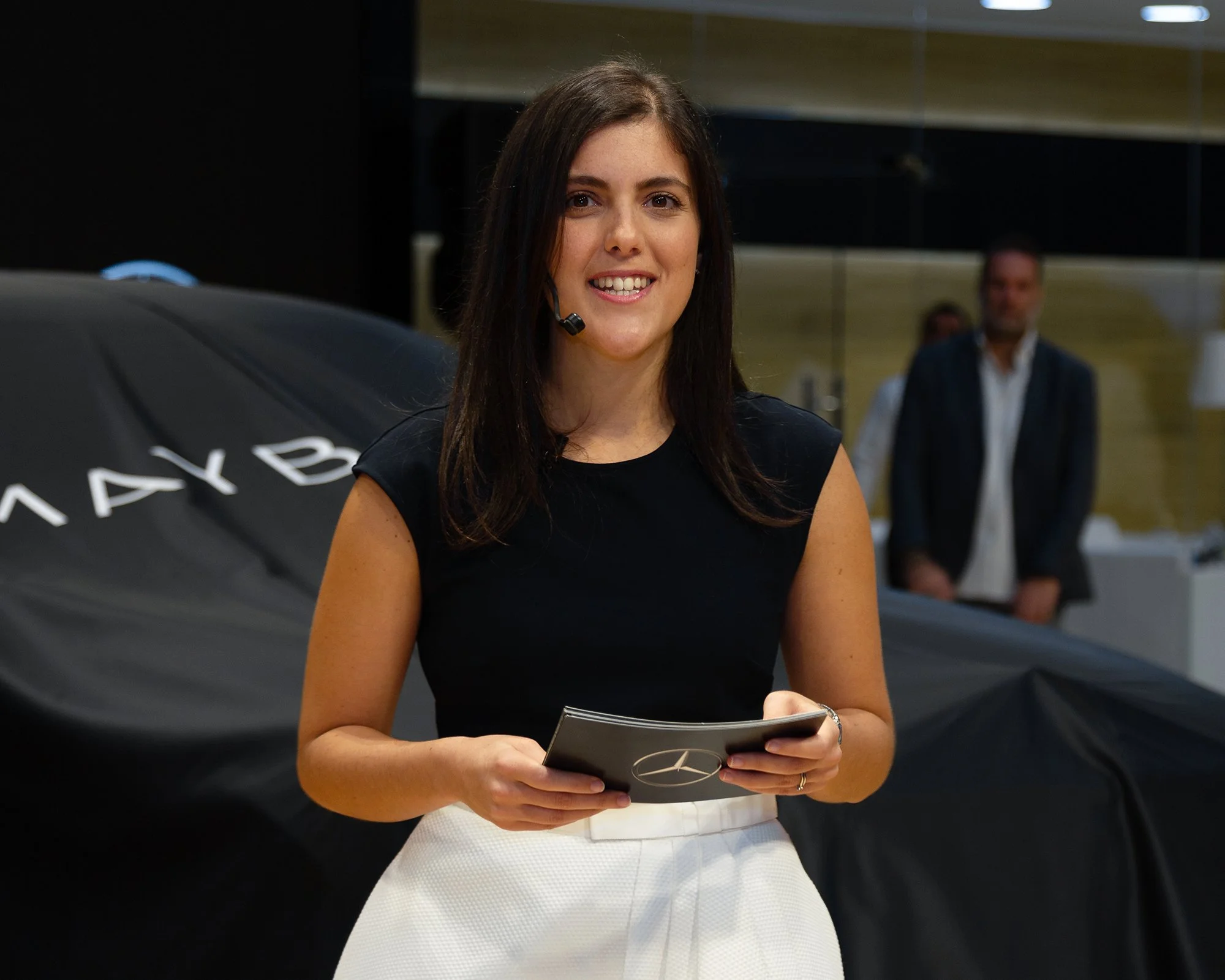 A smiling woman holding a booklet with Mercedes-Benz logo at an event, with a covered car and people in the background.