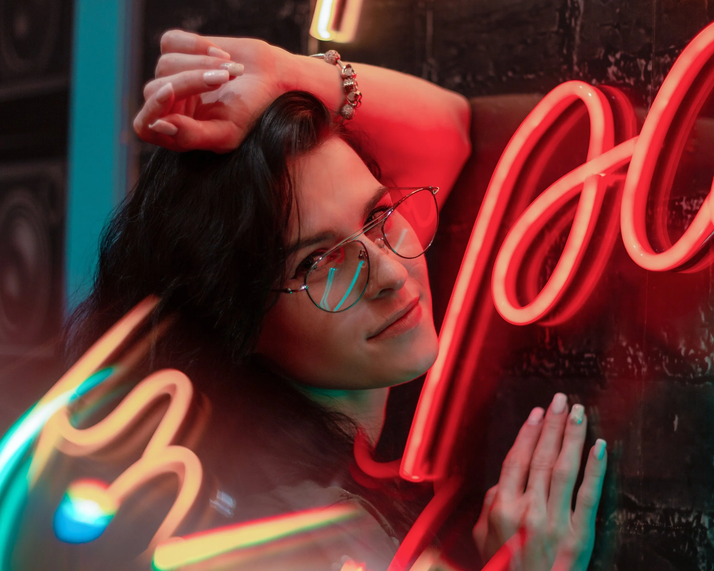 A woman with dark hair and glasses leaning against a wall with neon lights, smiling gently at the camera.