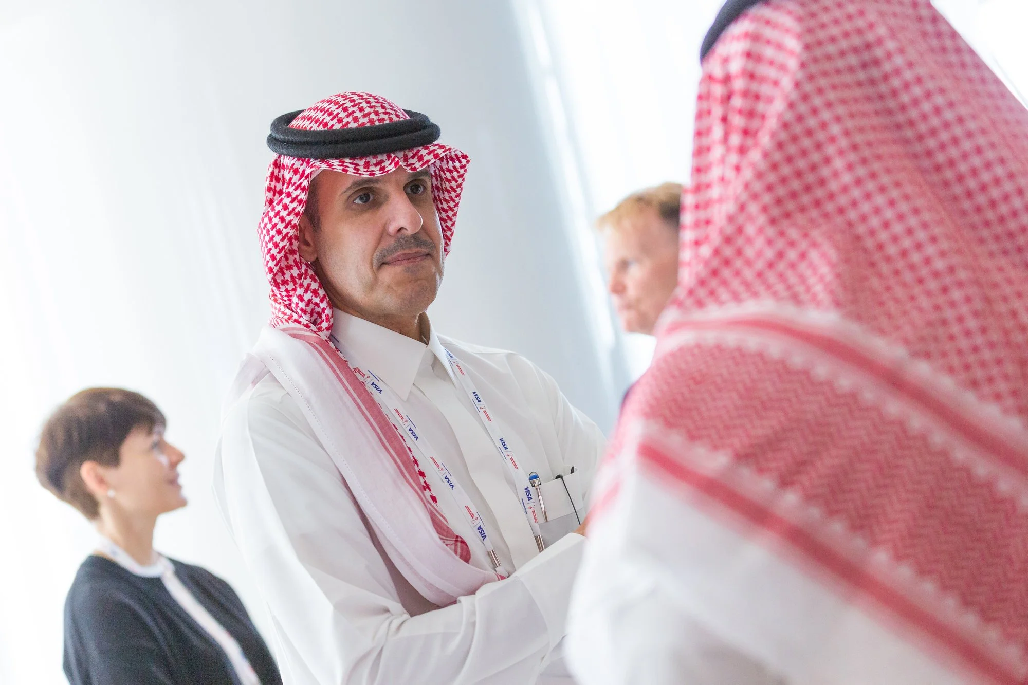 Man wearing a traditional Middle Eastern keffiyeh and white thobe engaged in conversation at a conference, with other attendees in the background.