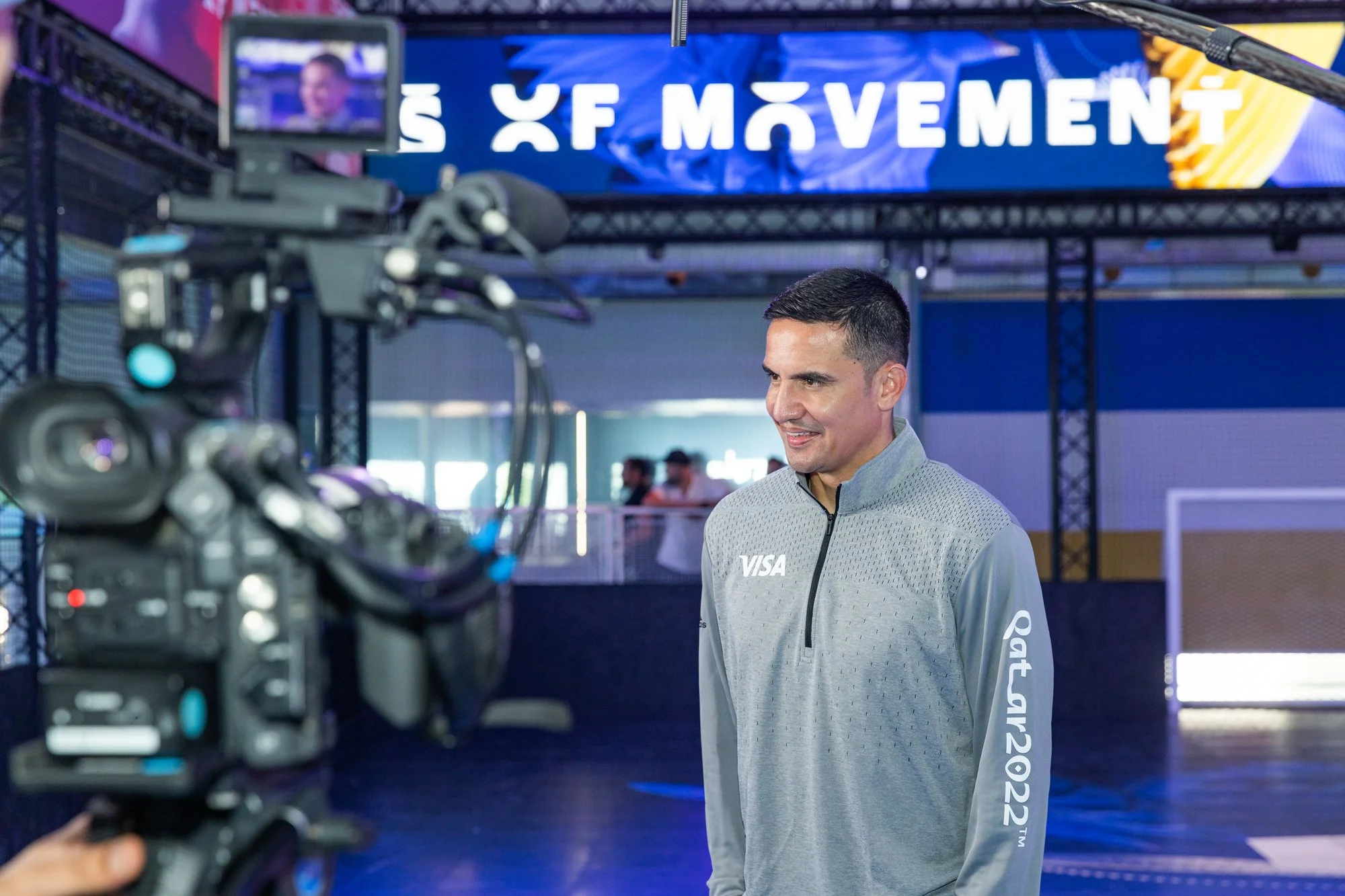 Man being interviewed on a sports court, wearing a gray athletic jacket with 'VISA' logo and 'Qatar 2022' on sleeve, with a video camera recording him, and a digital screen in the background displaying the words 'X X MOVEME...'