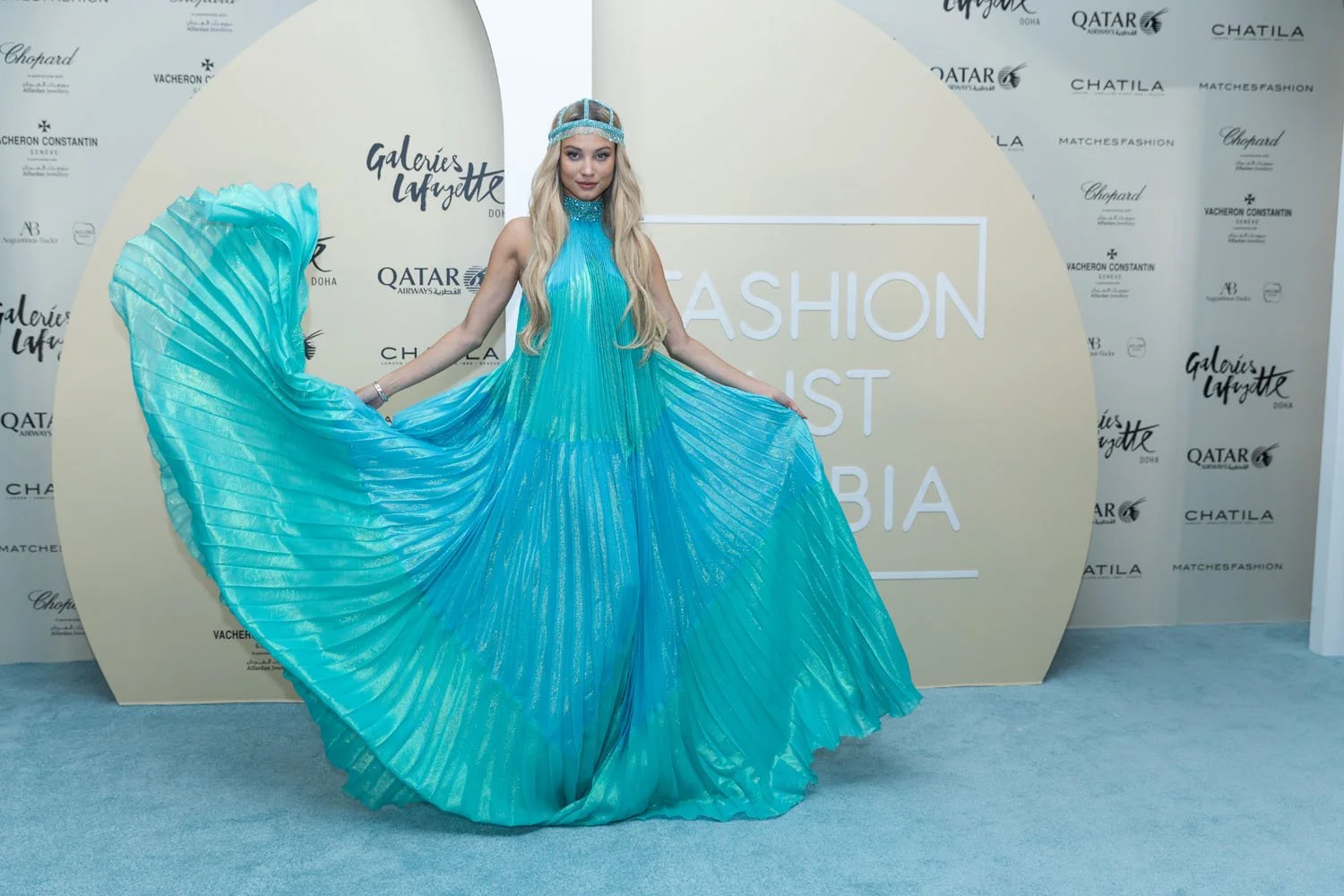 A woman in a flowing, shiny turquoise gown on a runway or event, holding the dress out to showcase its pleated design. The background features logos and text, including 'Galeries Lafayette Doha' and 'Fashion List Qatar'.