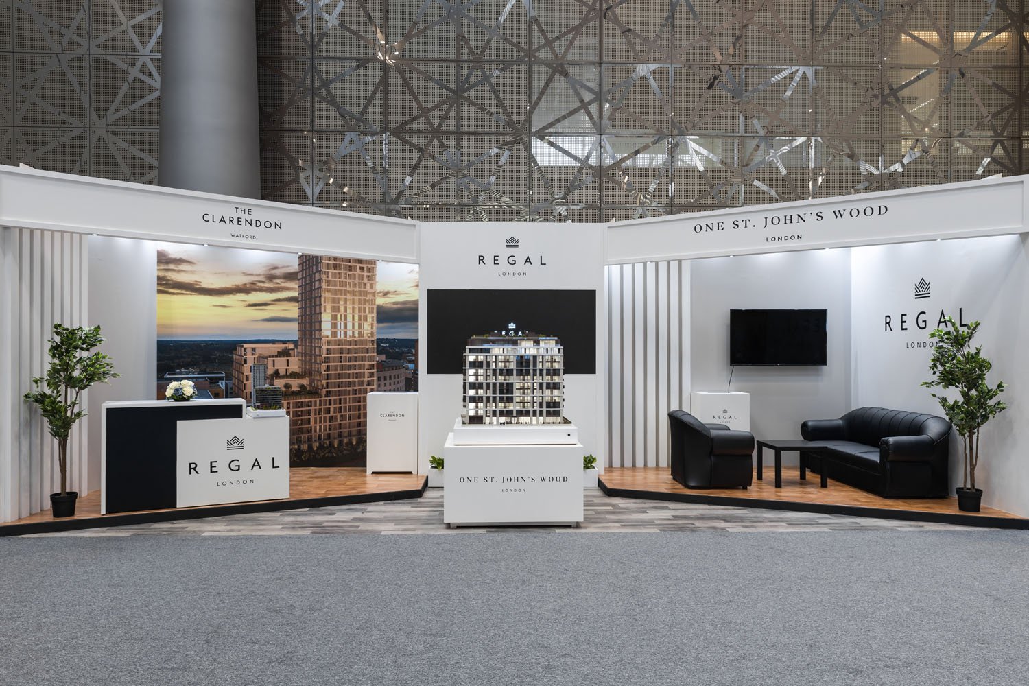 Exhibition booth for Regal London with a model of a residential building, black sofa, TV, and plants, featuring branding for The Clarendon, One St. John’s Wood, and Regal London.