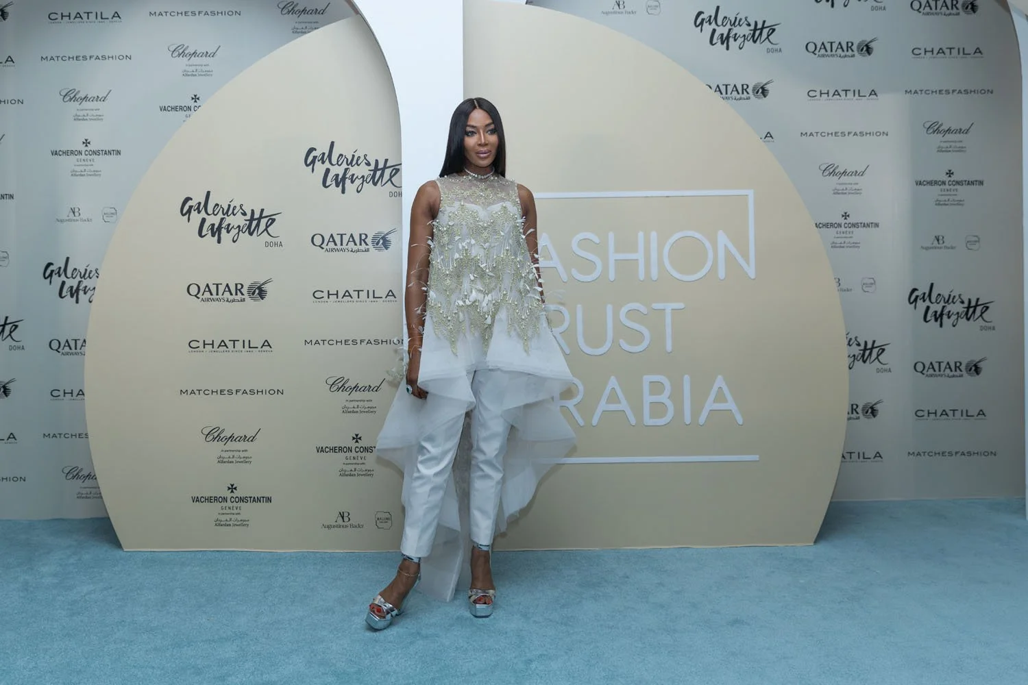 A woman in a white, embellished top with floral details and white pants stands on a blue carpet in front of a backdrop with logos and the words "Fashion Trust Arabia."