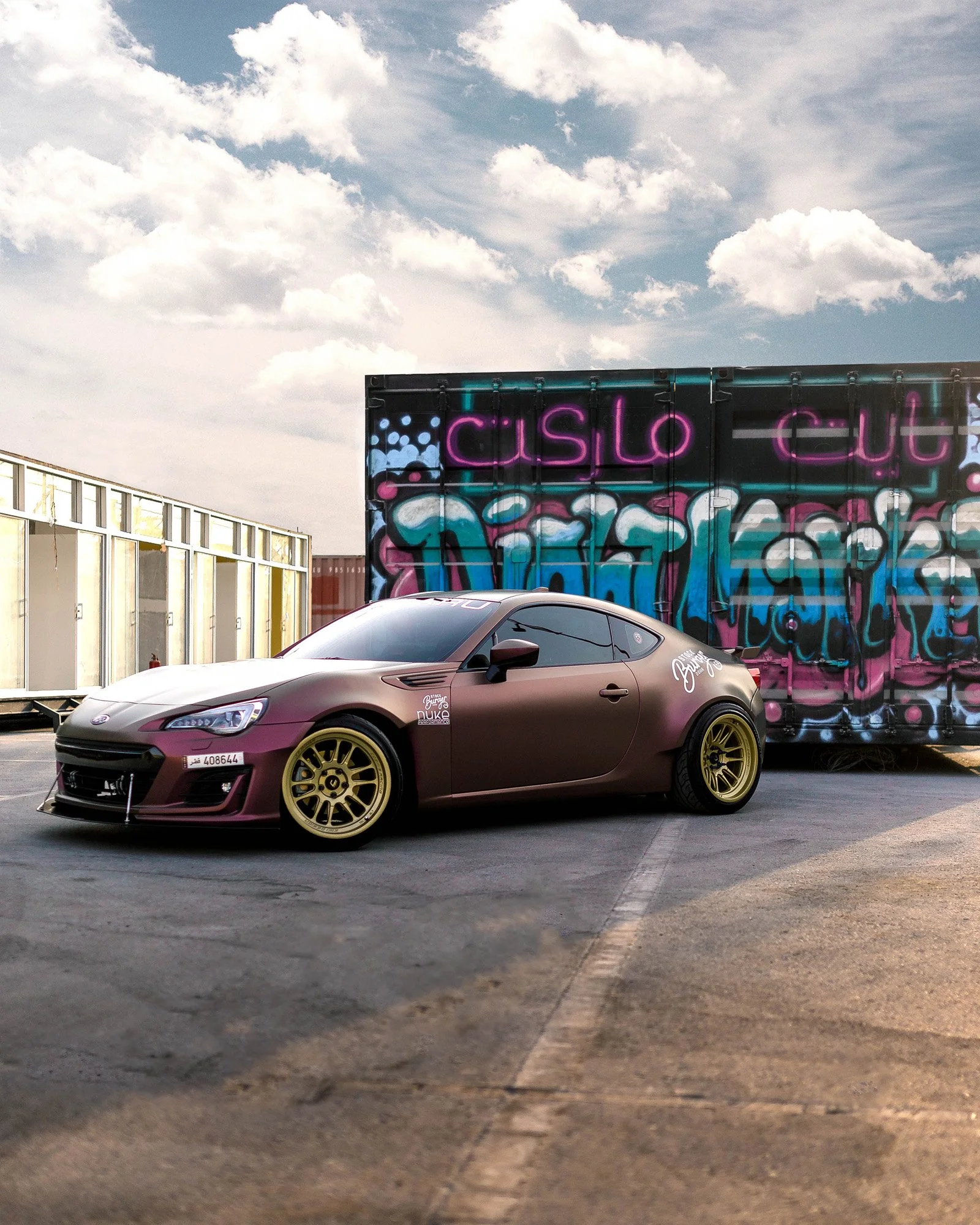 Gray sports car with yellow rims parked in front of colorful graffiti wall that reads 'CASLO' and 'DUKE'