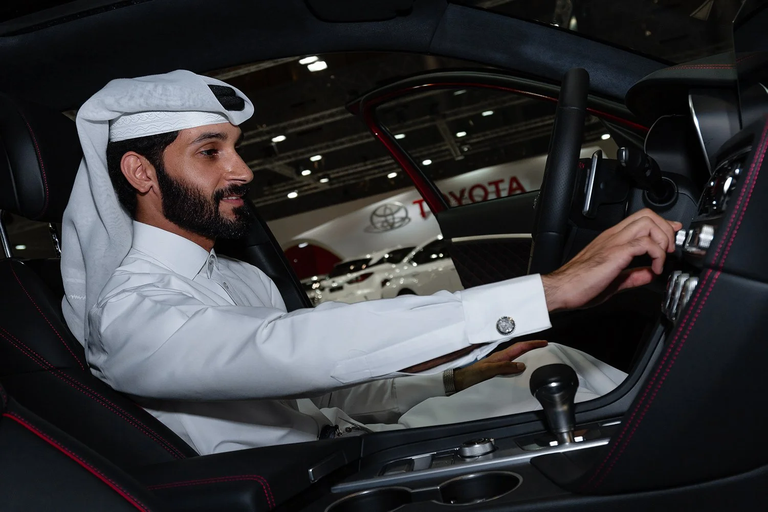 Man in traditional Middle Eastern clothing sitting in a car at an auto show, adjusting the controls on the dashboard.