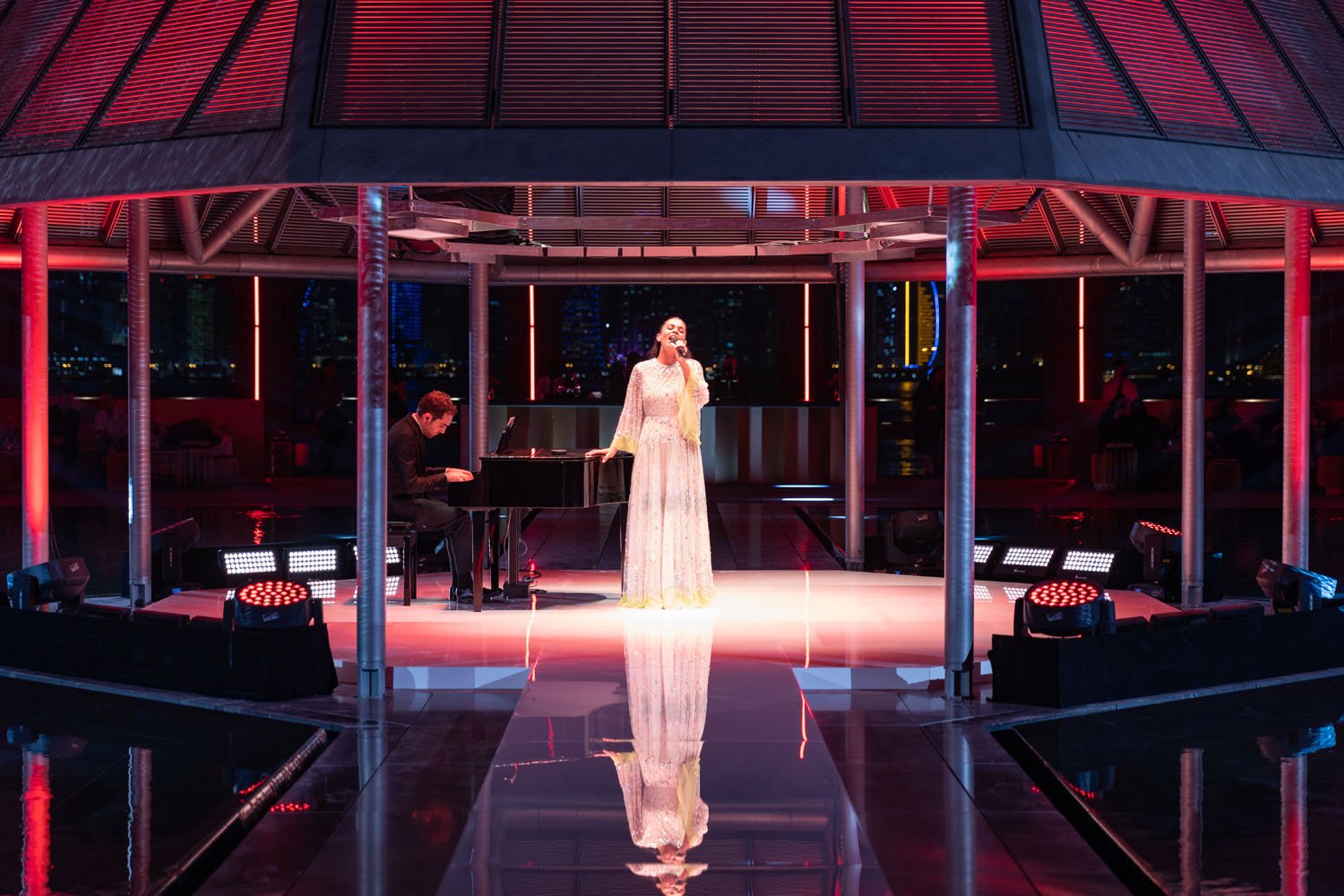 A woman in a long white dress singing into a microphone on a stage with a pianist accompanying her at night, with cityscape in the background and colorful stage lighting.