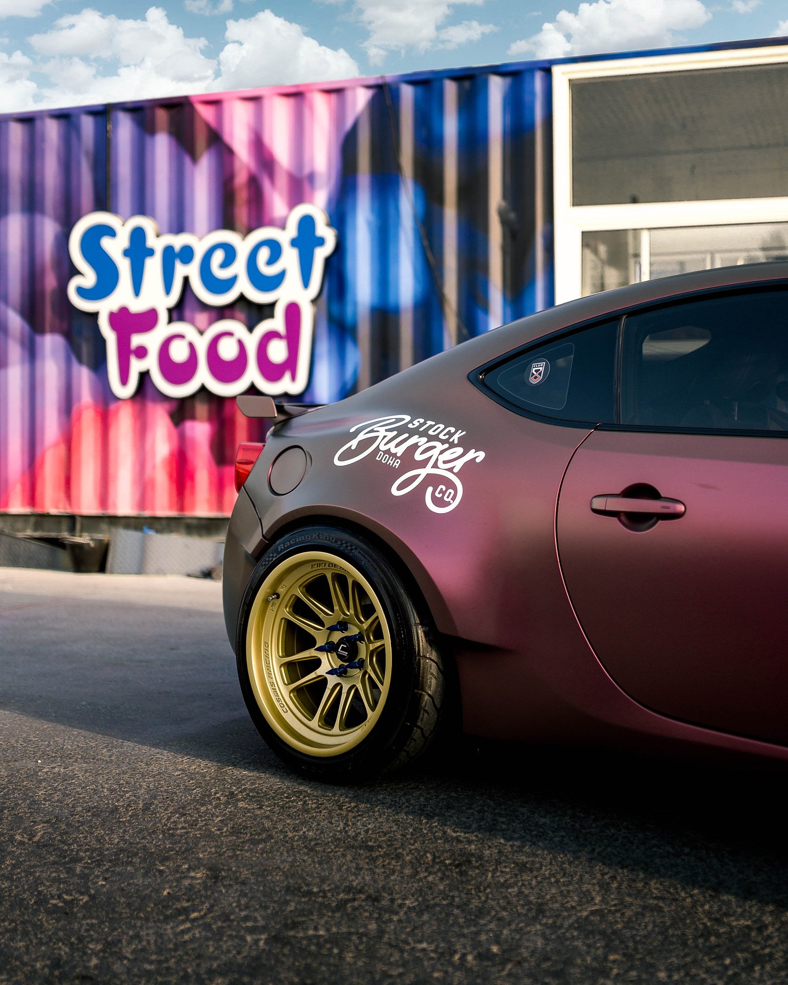 A matte purple sports car with gold wheels parked in front of a colorful mural that reads 'Street Food'.