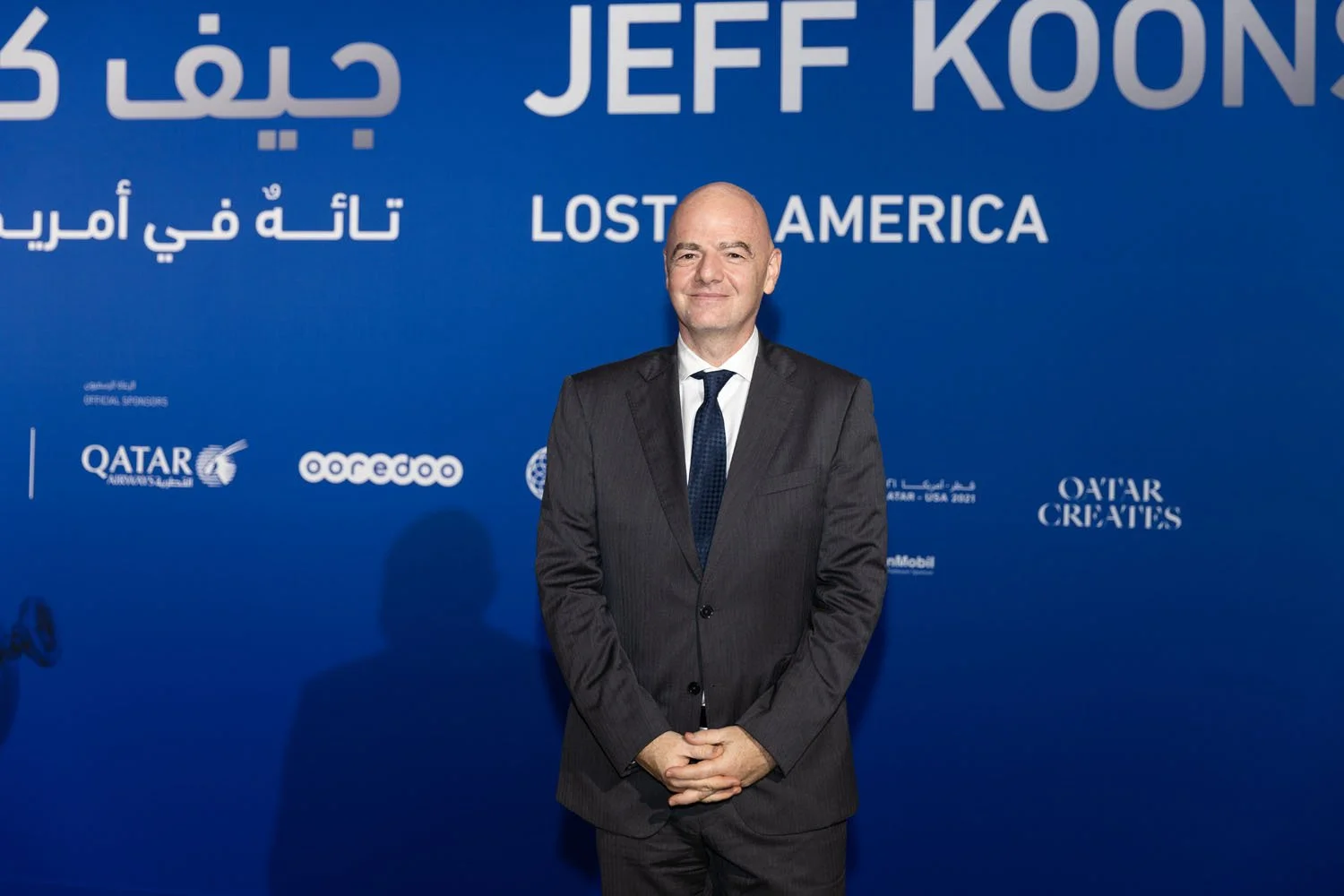 A man in a dark suit and tie standing in front of a blue backdrop with white text and logos, including 'Jeff Koons' and 'Lost in America'.